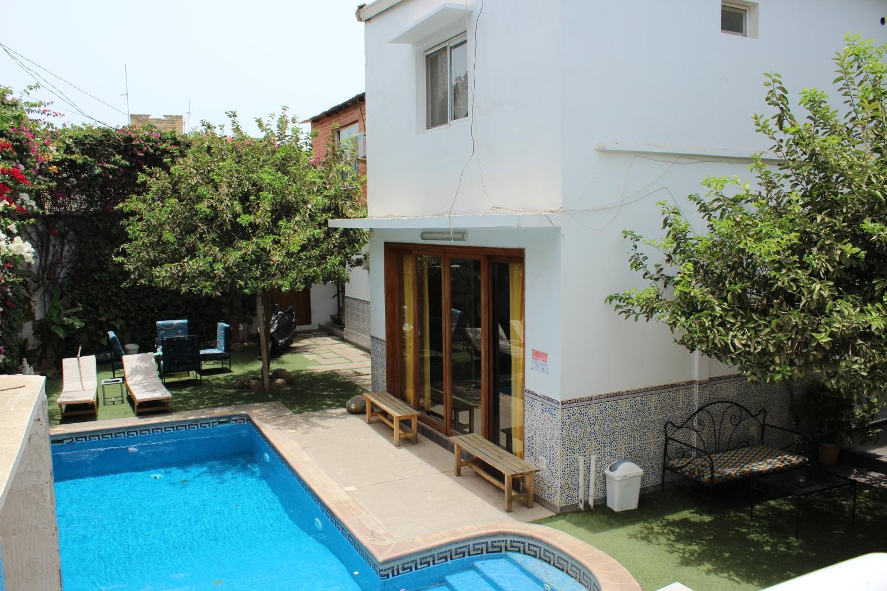 A swimming pool with lounge chairs, a wooden bench, and a covered patio area with a tiled wall and a potted plant.