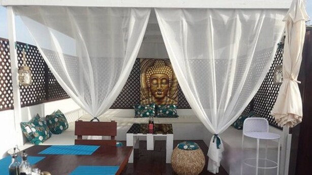 A room with a wooden floor, a dining table with blue placemats, a white chair, a large Buddha statue, and a white canopy. - Casa Mara Guest House (Dakar)