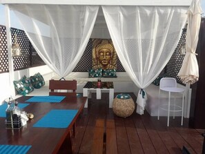 A room with a wooden floor, a dining table with blue placemats, a white chair, a large Buddha statue, and a white canopy. - Casa Mara Guest House (Dakar)
