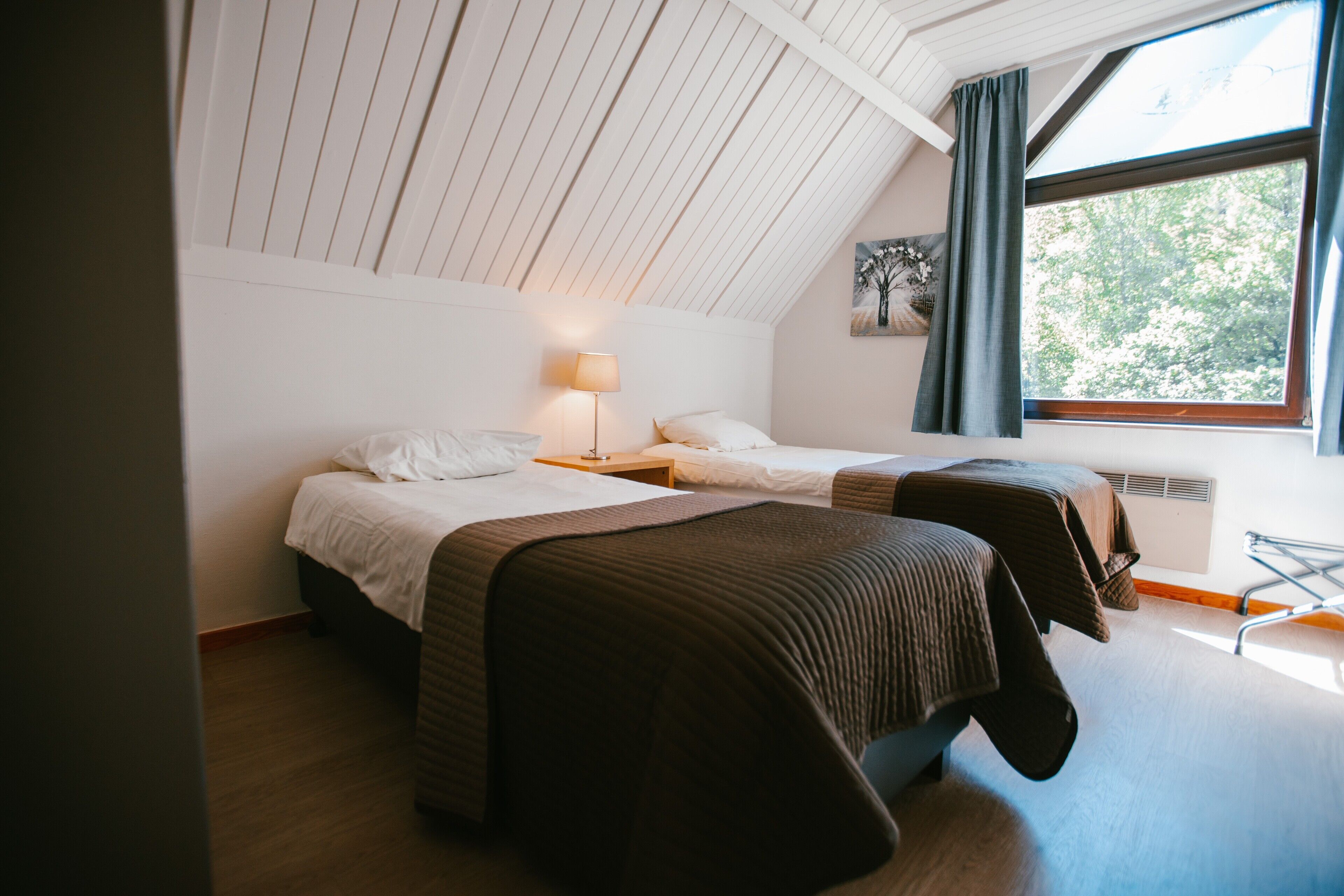 A room with two beds, a standing lamp, a window with curtains, and a painting on the wall.