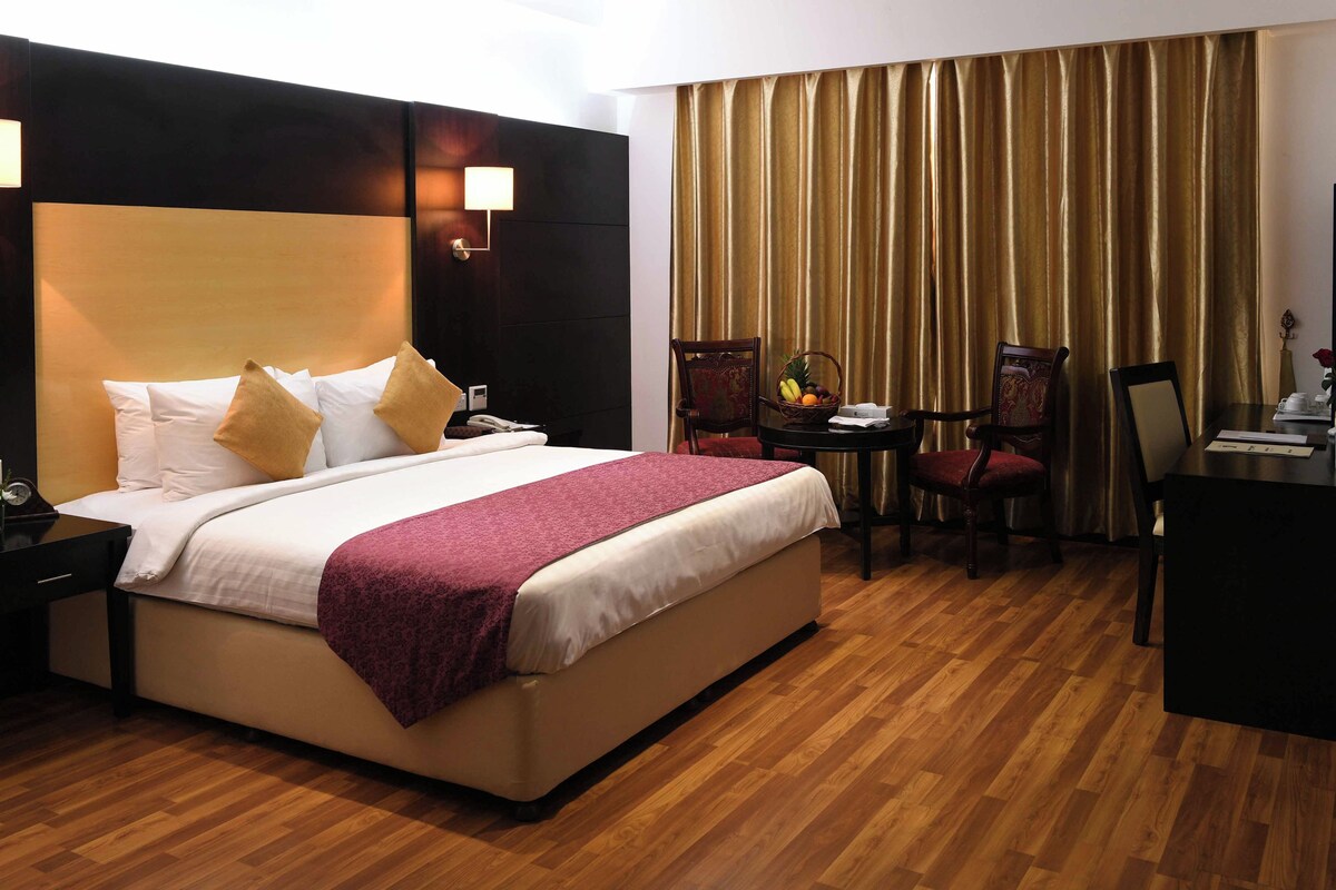 A hotel room with a large bed, bedside tables, a desk with a chair, a small table with fruit, and a large window with curtains.