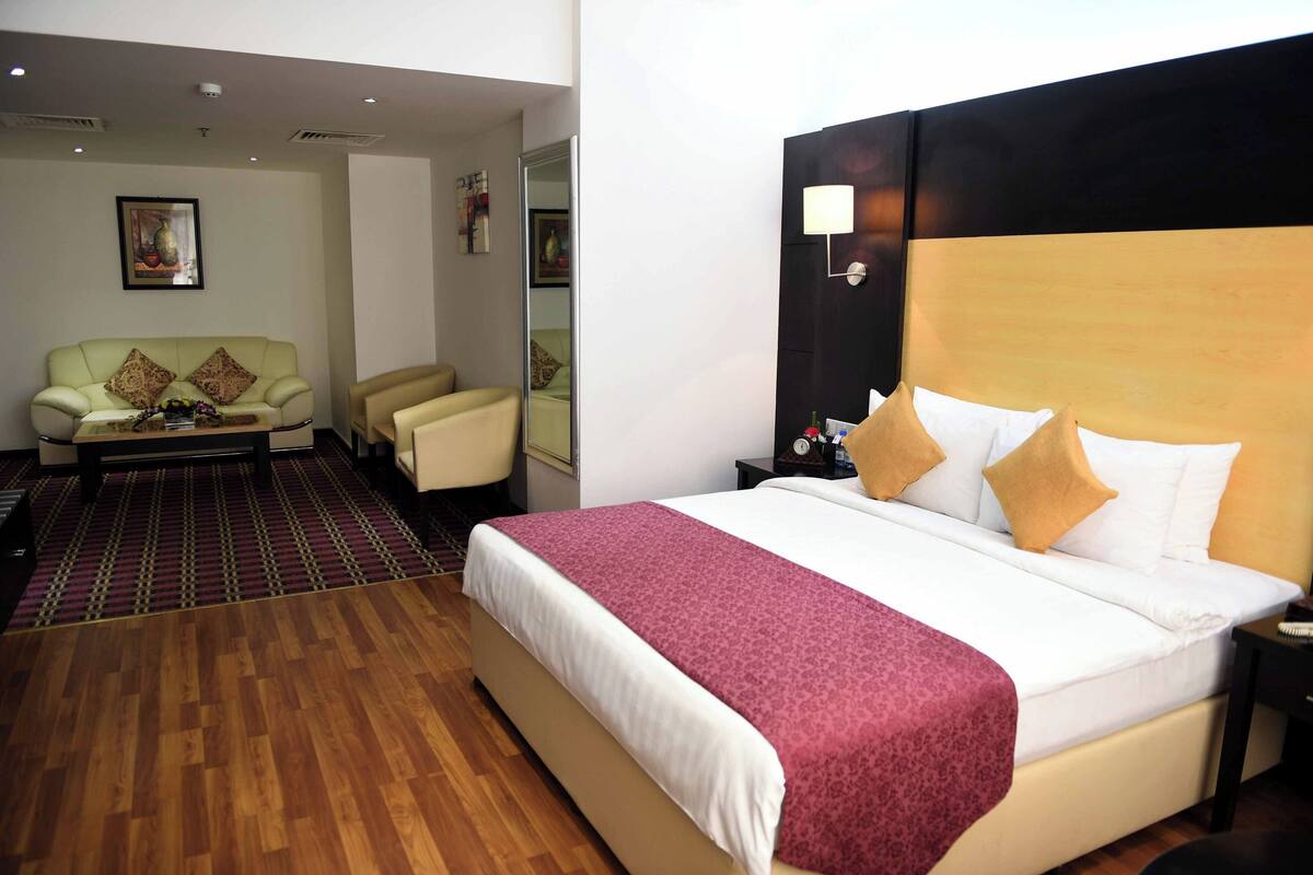 Una habitación de hotel con una cama grande, una zona de estar con sofá y sillones, piso de madera y un espejo.