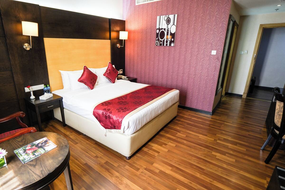Habitación de hotel con una cama grande, dos mesitas de noche, suelo de madera, pared roja y una pequeña mesa con una revista y un florero.