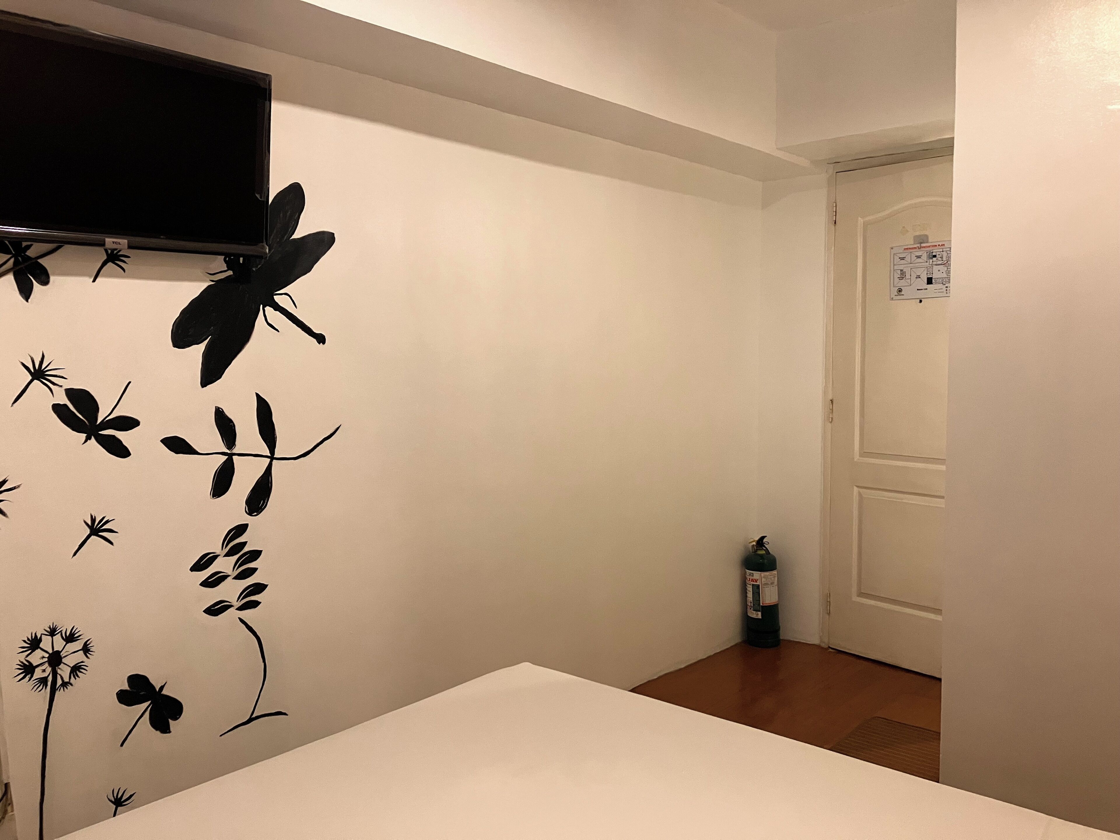 Een kamer met een witte muur, een wandgemonteerde flatscreen-tv, een deur met een bordje en een flesje op de vloer.