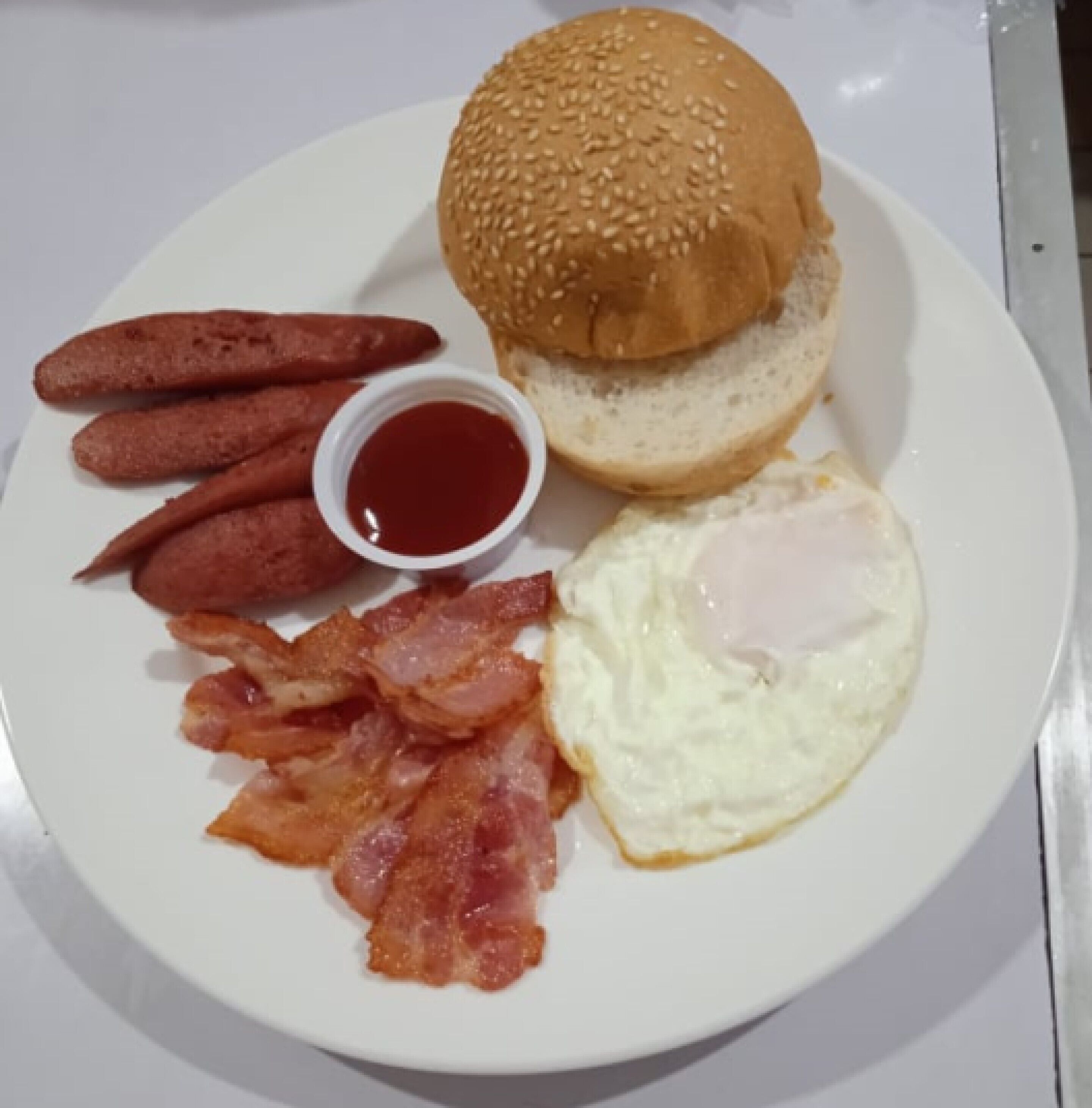 A plate with a hamburger, bacon, eggs, and a small container of ketchup.