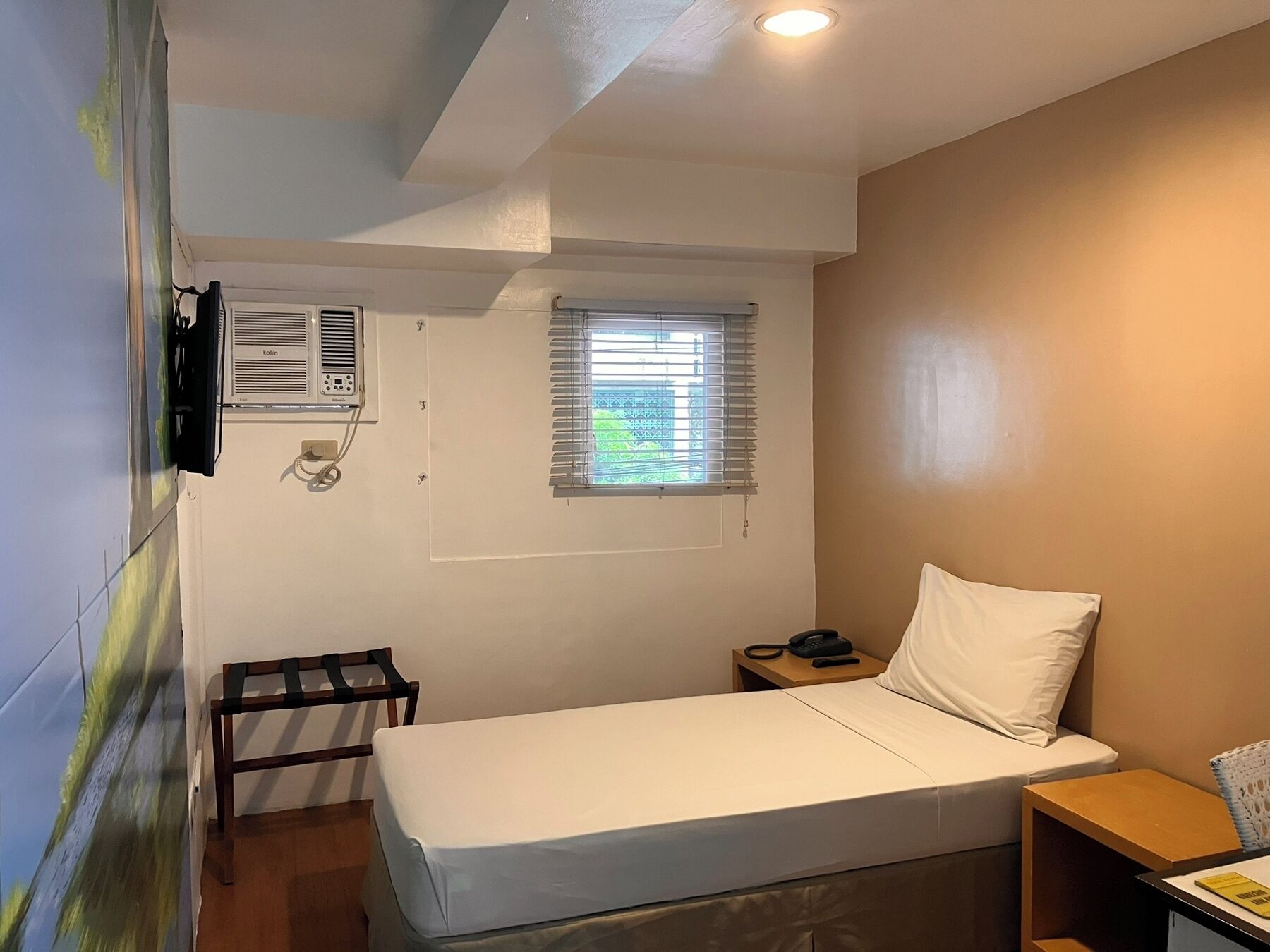 Ein kleines Zimmer mit einem Einzelbett, einem Fenster, einer Wand-Klimaanlage, einem Nachttisch mit Lampe und einem Sessel.