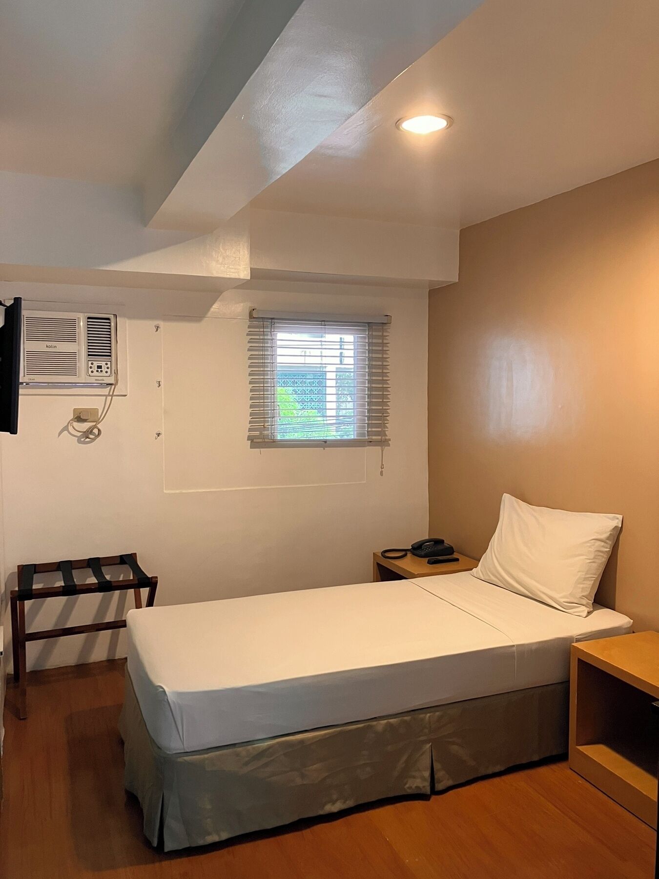 Une petite chambre avec un lit simple, un plancher en bois, une fenêtre avec des stores, un climatiseur fixé au mur et une table de chevet avec un téléphone.