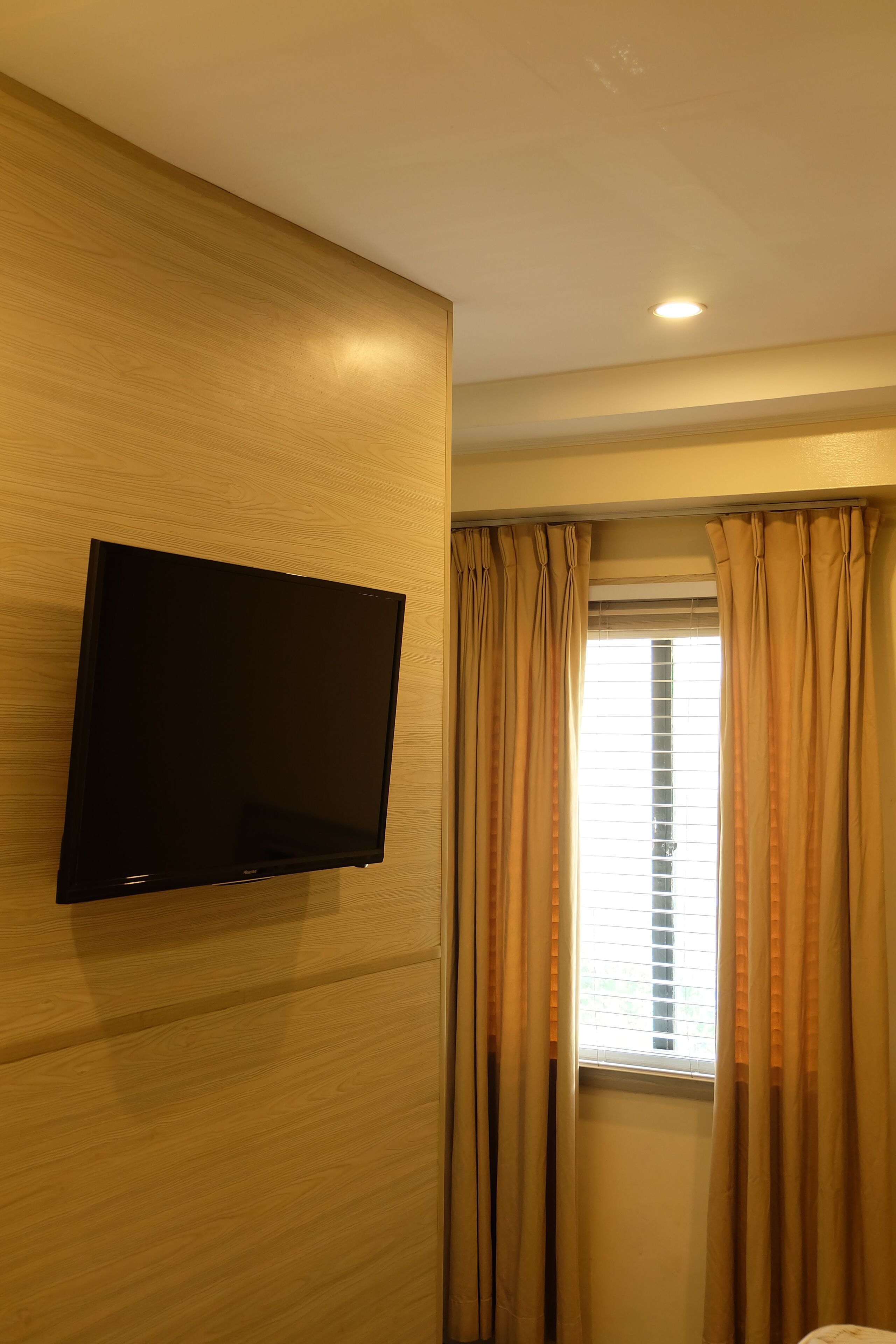 A room with a mounted flat-screen TV on a wooden wall, beige curtains, and a window with blinds.