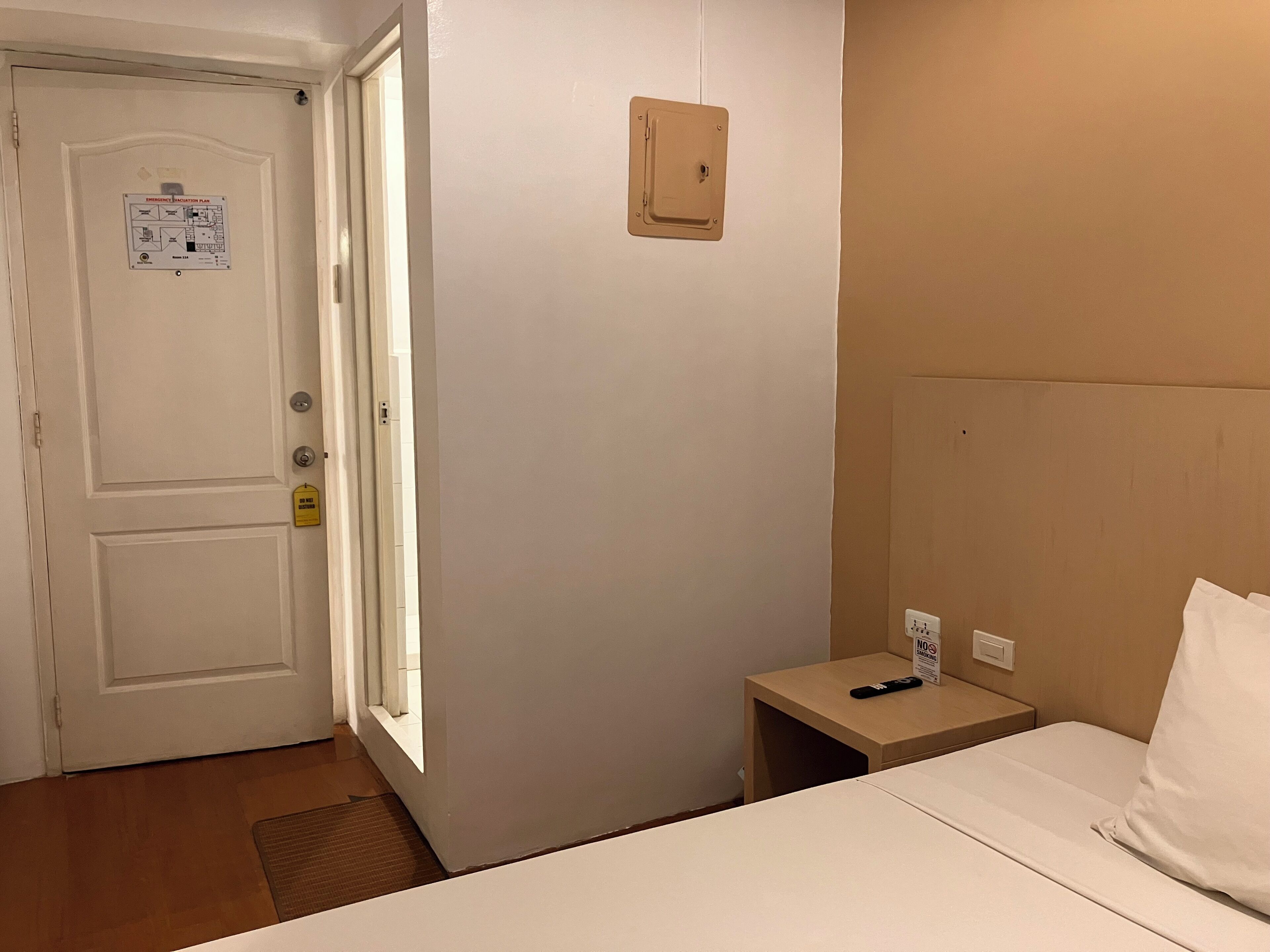 A hotel room with a bed, a bedside table with a remote control, a wall-mounted light switch, and a door with an emergency evacuation plan poster.