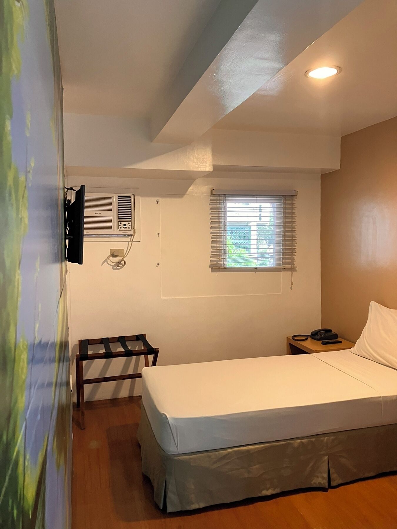 Ein Schlafzimmer mit einem Bett, einem kleinen Tisch mit Telefon, einem Fenster mit Jalousien, einer Klimaanlage und einem an der Wand montierten Fernseher.