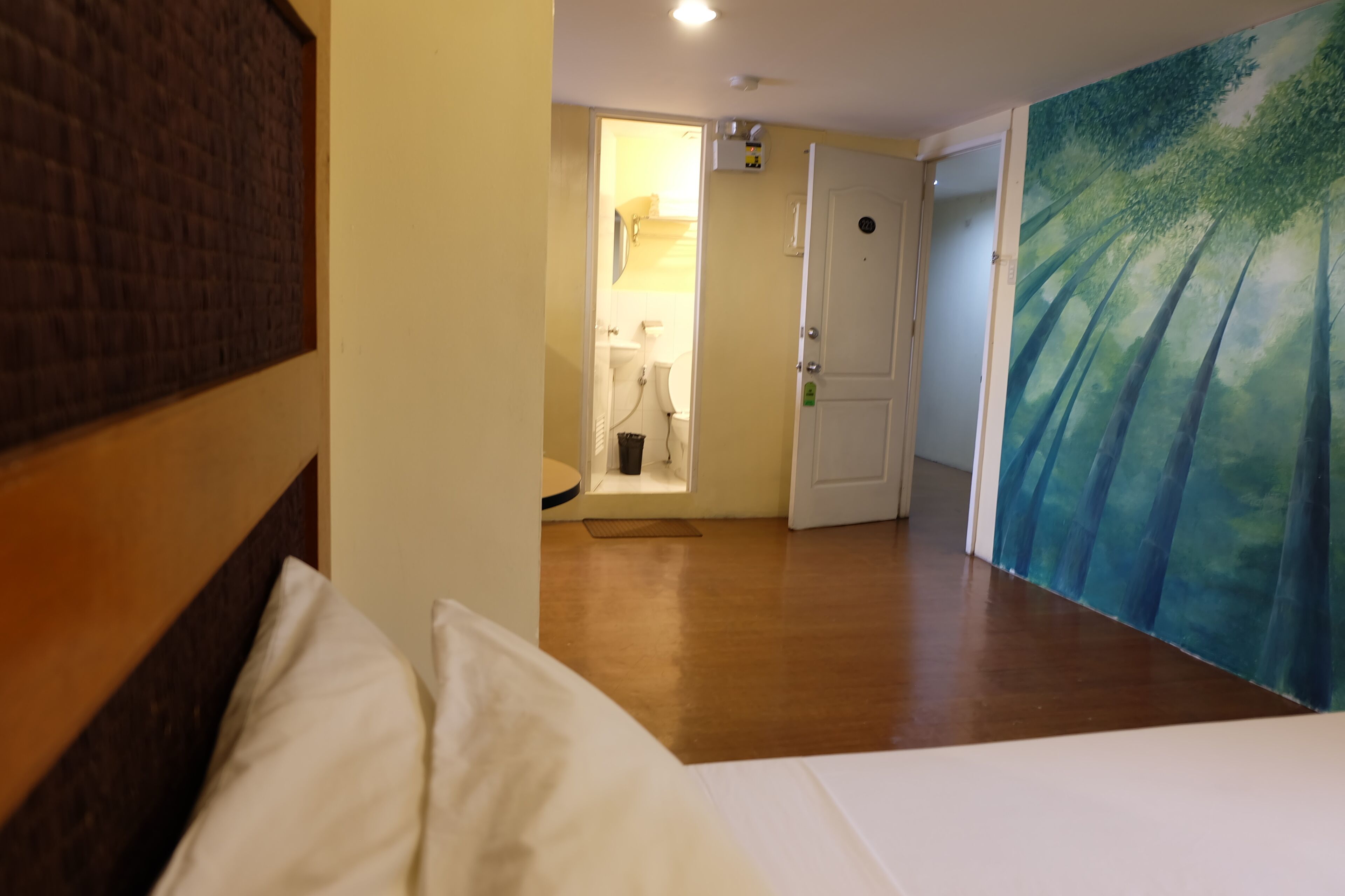 Une chambre d’hôtel avec une tête de lit en bois, un lit avec une literie blanche, une salle de bain avec des toilettes et un lavabo, et un tableau représentant une forêt accroché au mur.