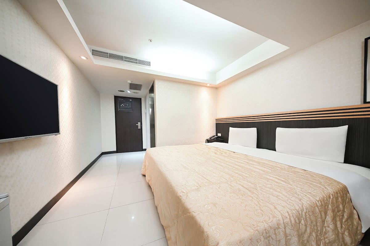 A hotel room with a bed, a flat-screen TV, a desk with a phone, and a door with a digital display.
