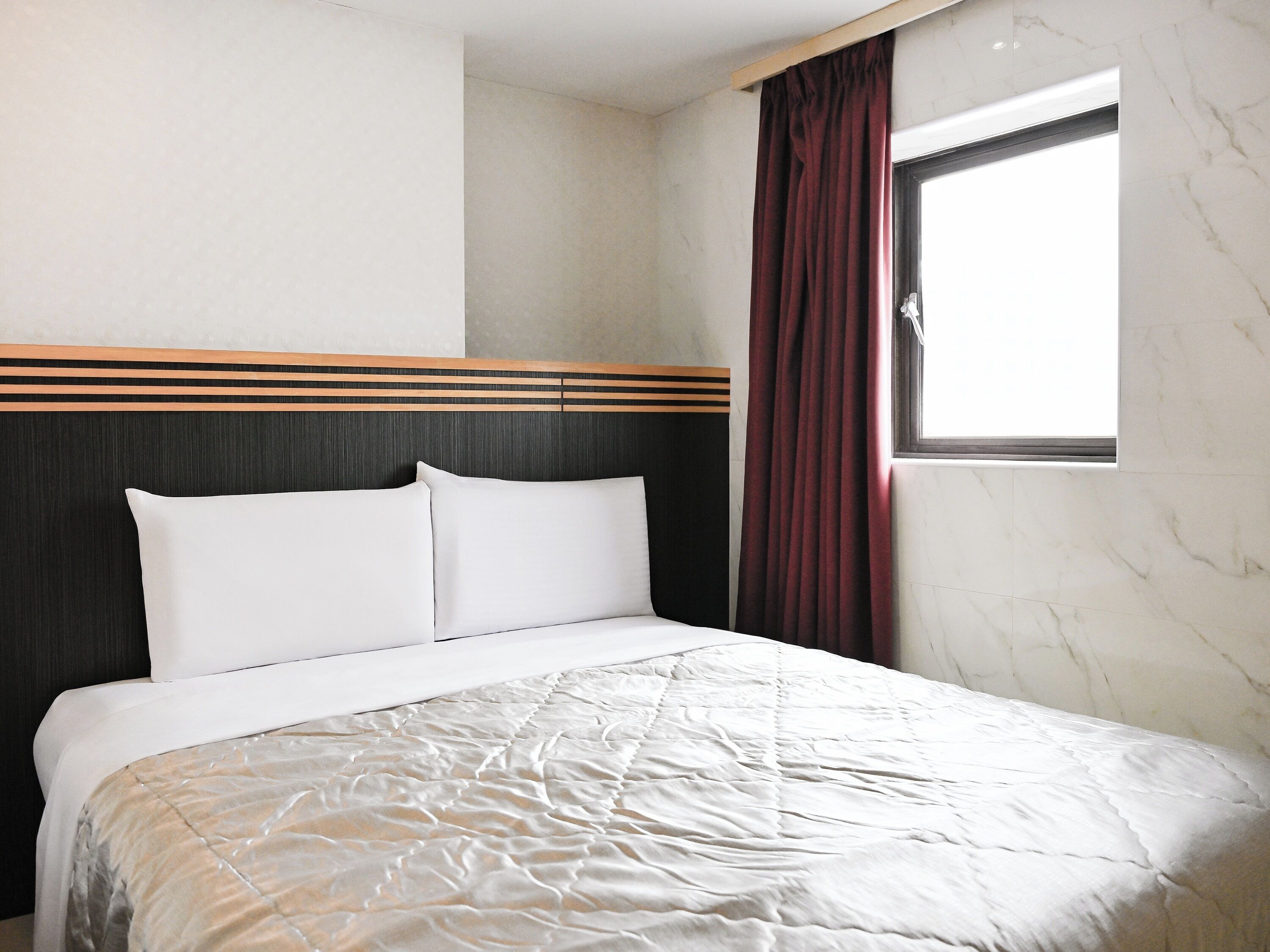 A hotel room with a bed, a window with curtains, a headboard with horizontal stripes, and a marble wall.