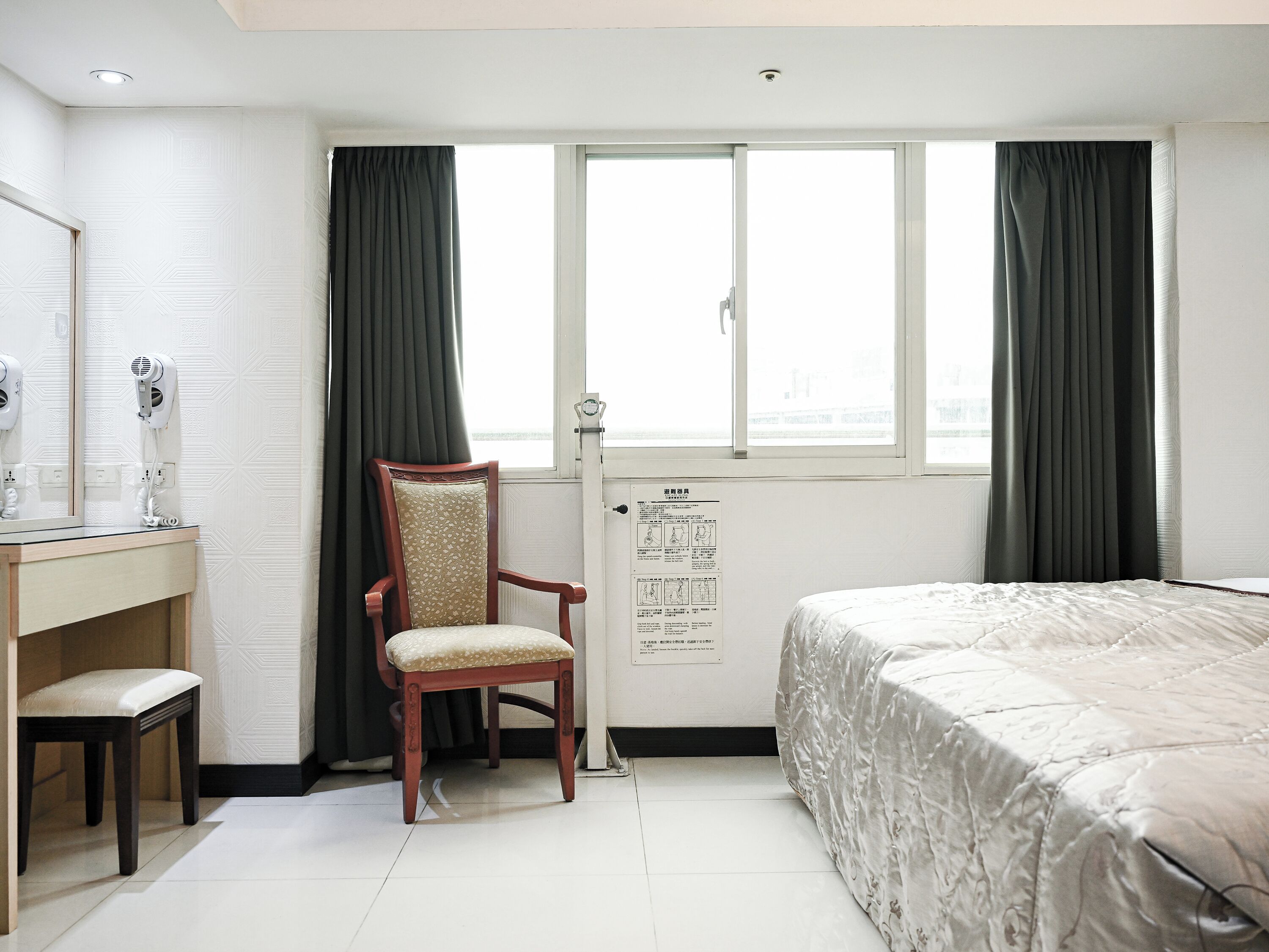 Habitación de hotel con cama, sillón, escritorio con secador de pelo, ventana con cortinas y puerta con un cartel.