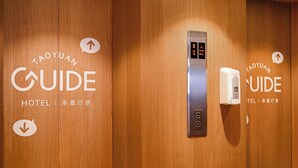 Et hotelinteriør med træpaneler, en digital elevatorvisning og en håndspritdispenser.