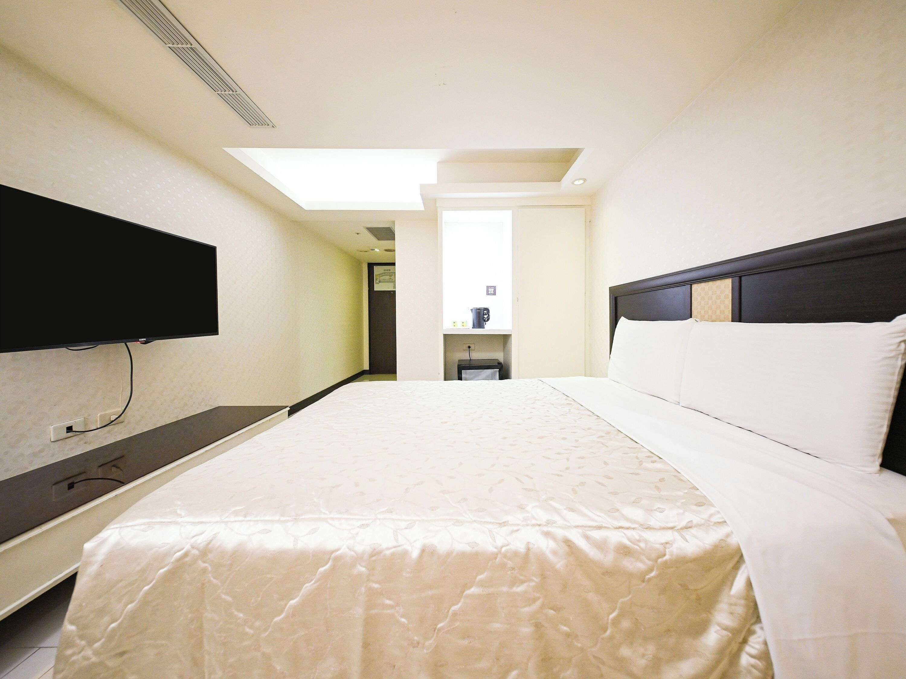 A hotel room with a bed, a flat-screen TV mounted on the wall, a desk, and a window providing natural light.