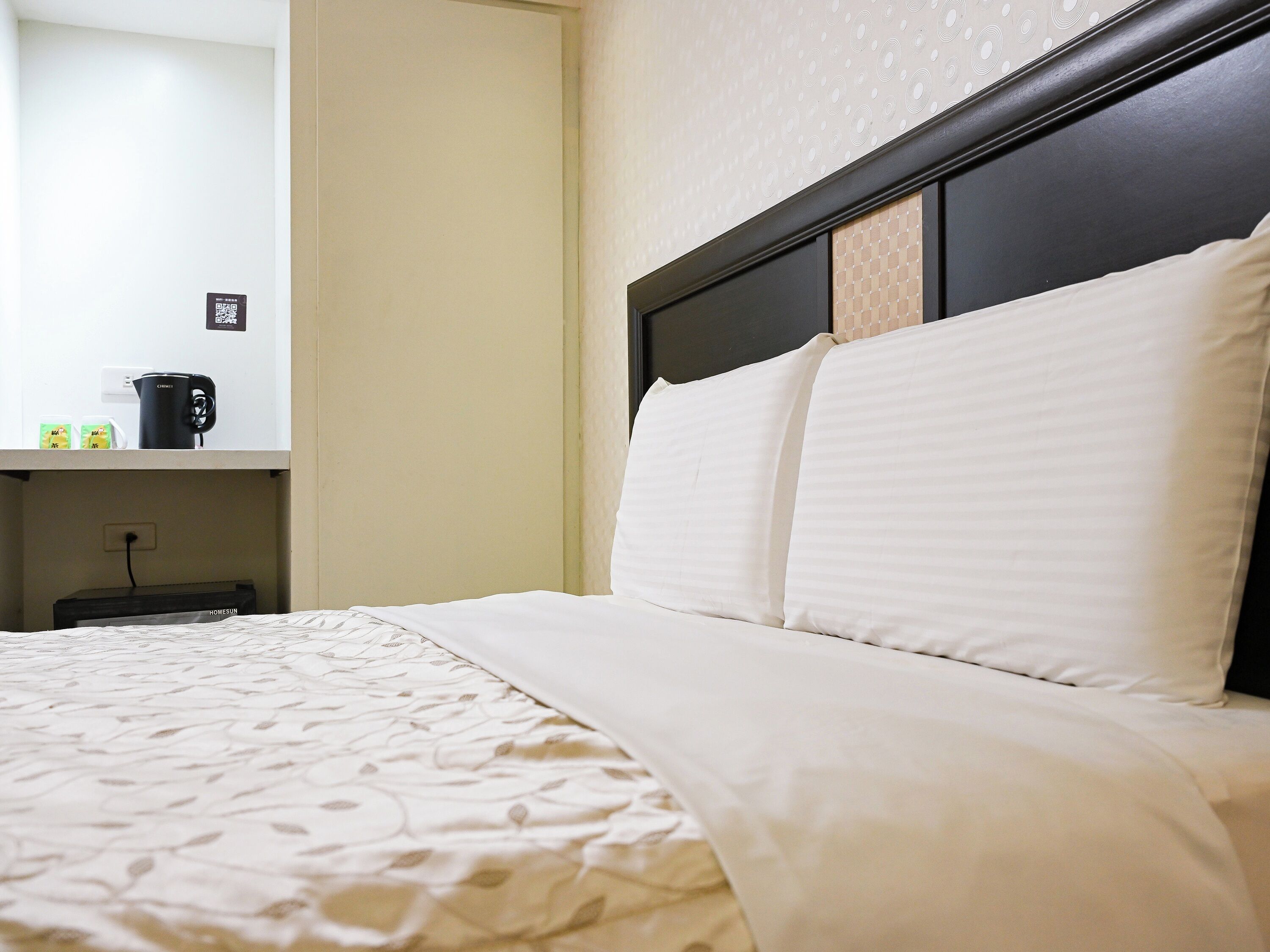 A neatly made bed with a patterned bedspread, a bedside table with a black mug, and a wall with a circular pattern.