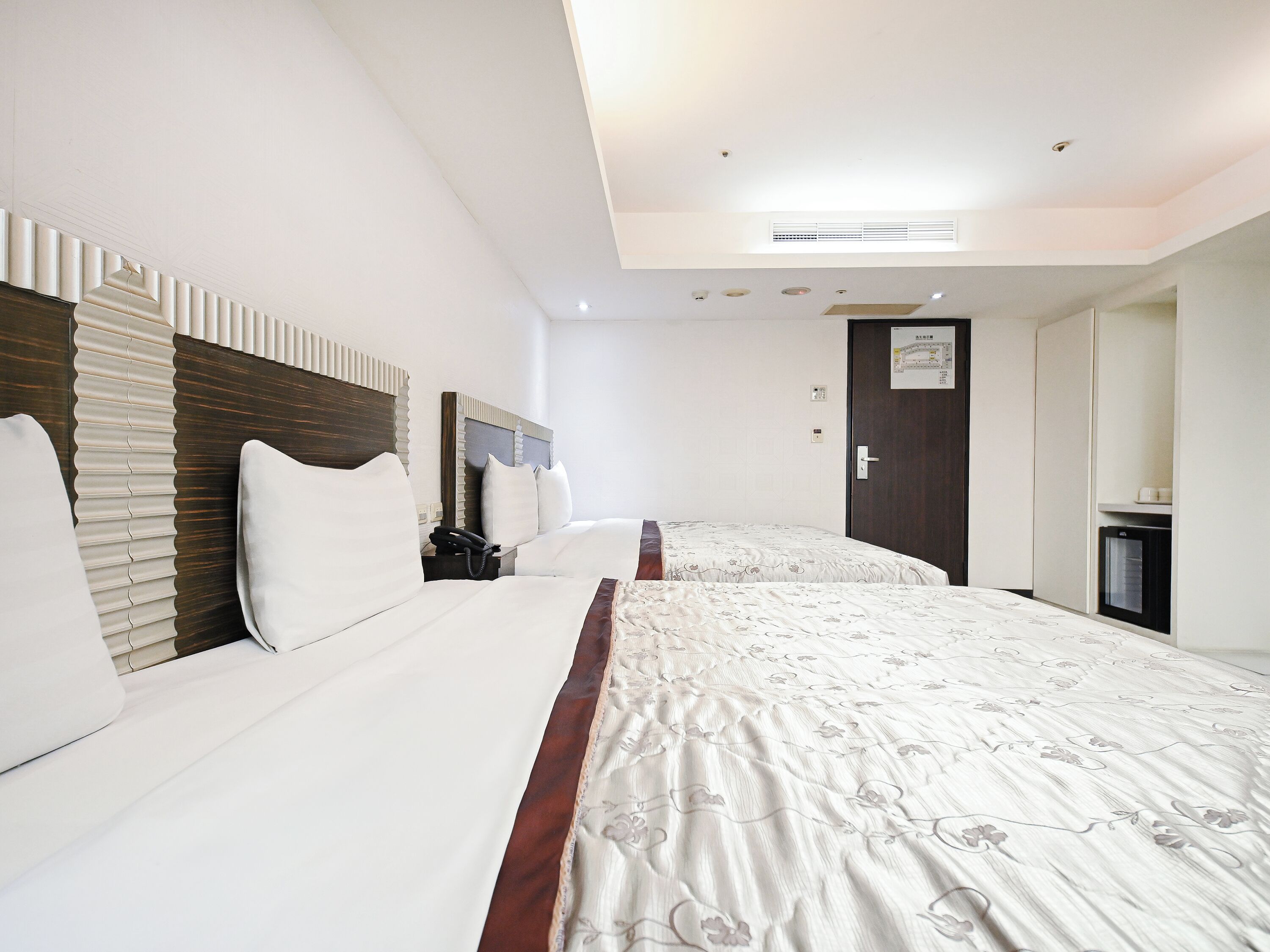 A hotel room with two beds, a wooden headboard, a bedside table with a phone, a door with a map, and a small kitchenette.