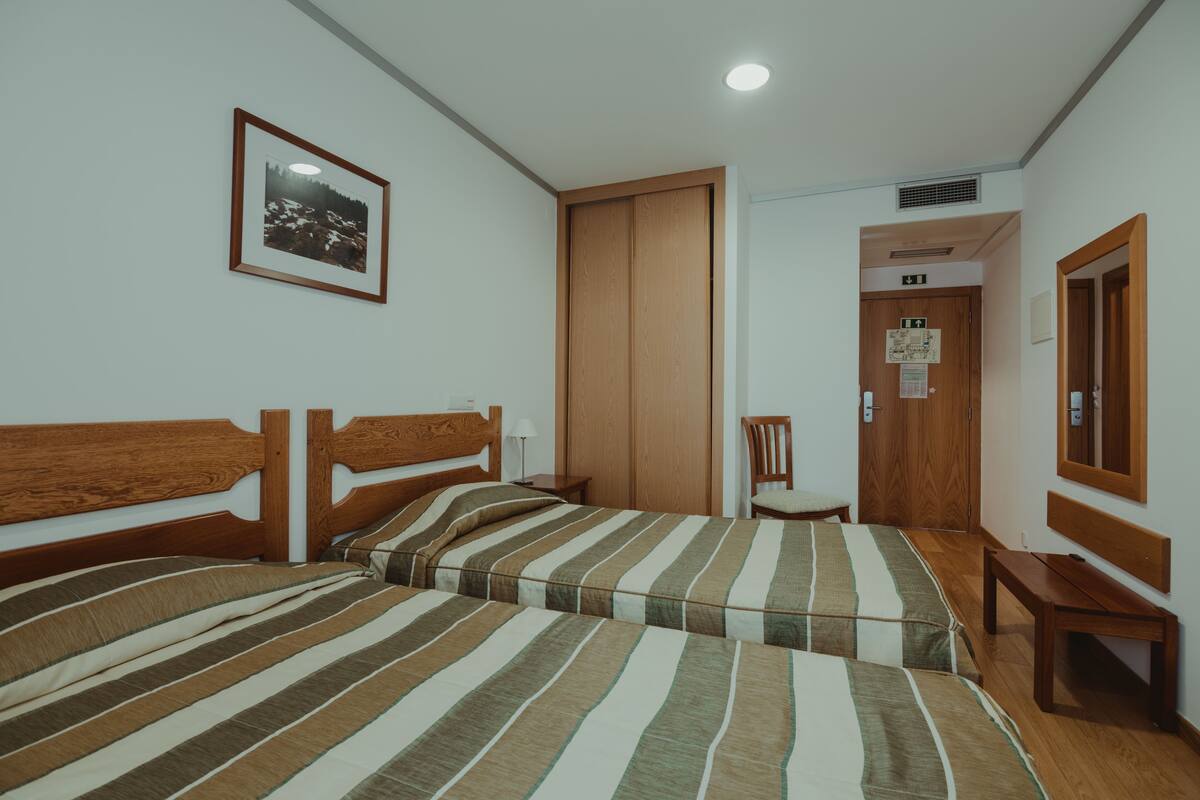 A hotel room with two beds, wooden headboards, a framed picture on the wall, a wooden chair, a small table, and a door with an emergency exit sign.
