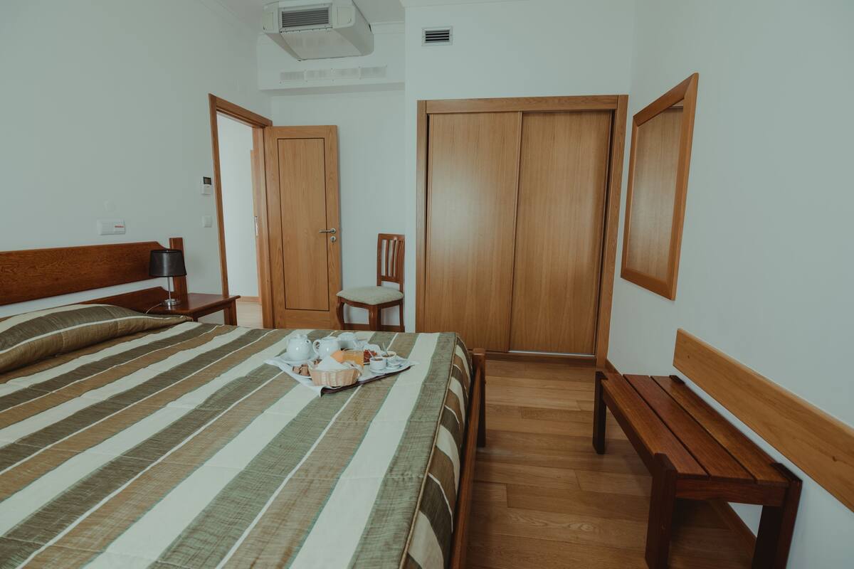 A bedroom with a bed, bedside table, chair, wooden wardrobe, and a bench.