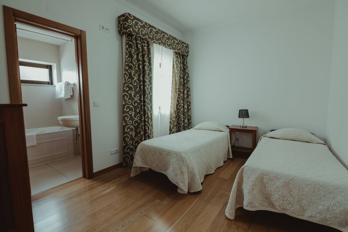 A bedroom with two beds, a nightstand, a window with curtains, a bathroom with a sink and bathtub, and wooden flooring.