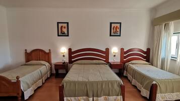 A hotel room with three single beds, each with a wooden headboard and a bedside table with a lamp.
