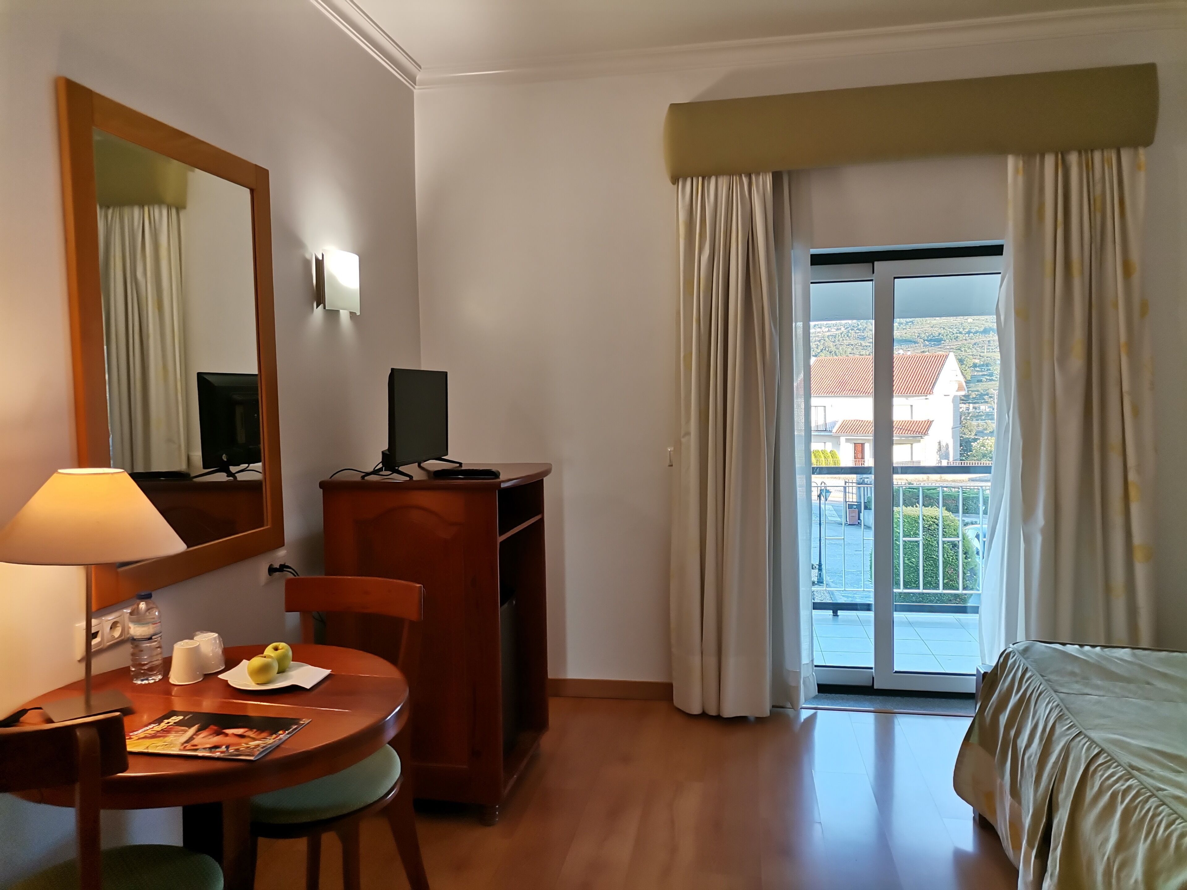Une chambre d’hôtel avec un bureau en bois, une chaise, un lit, un grand miroir, un téléviseur posé sur une commode en bois, et une vue sur un immeuble à travers une porte vitrée.
