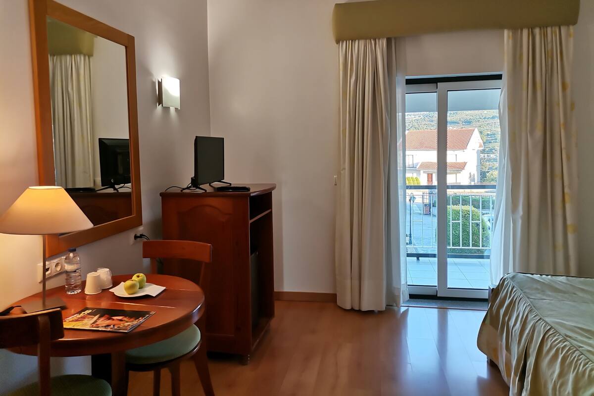 Ein Hotelzimmer mit einem Holztisch, einem Stuhl, einem Bett, einem großen Spiegel, einem Fernseher auf einer Holzeinrichtung und Blick auf ein Gebäude durch eine Glastür.