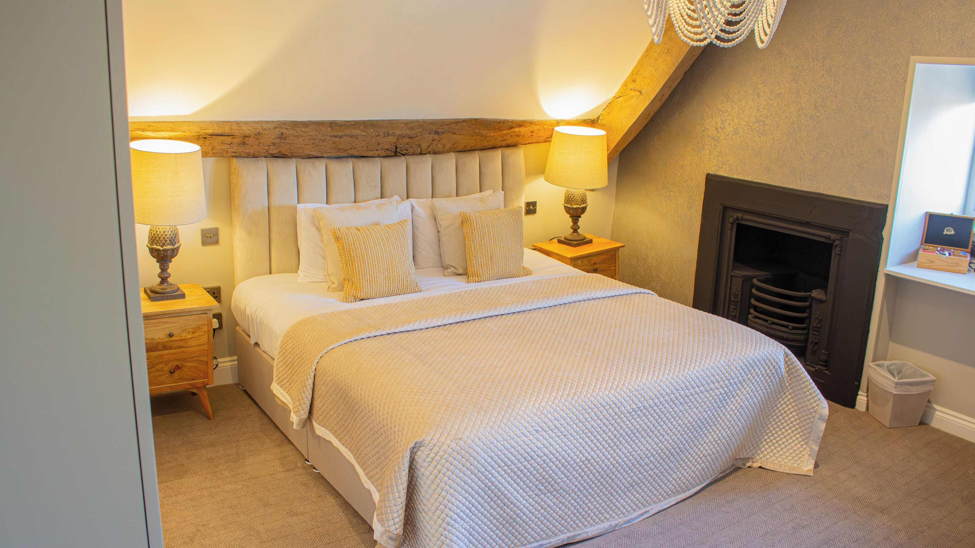 A bedroom with a large bed, two bedside lamps, a wooden headboard, a fireplace, and a small wooden shelf.