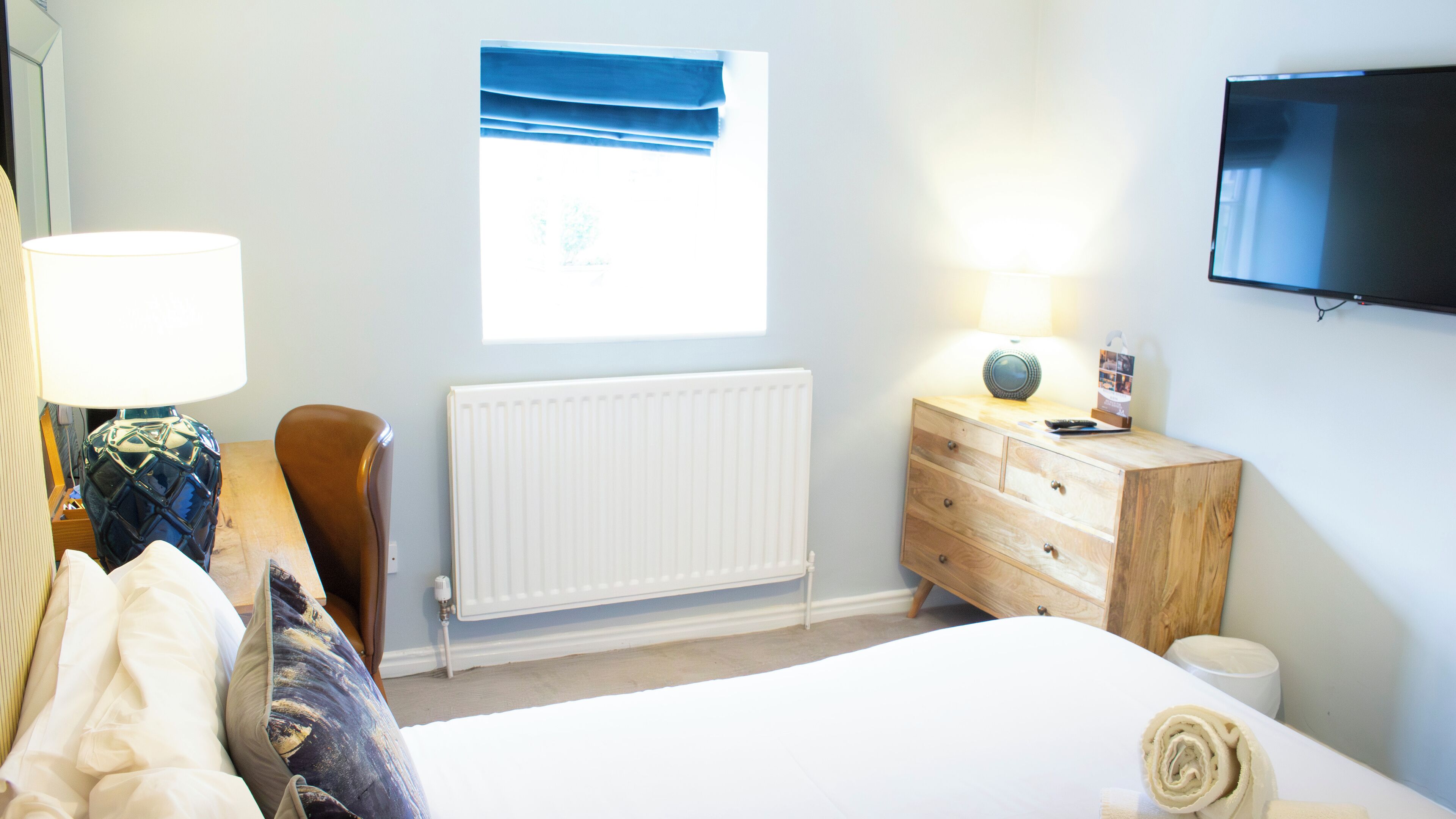 A bedroom with a bed, a dresser, a lamp, a radiator, a window with blinds, and a TV mounted on the wall.