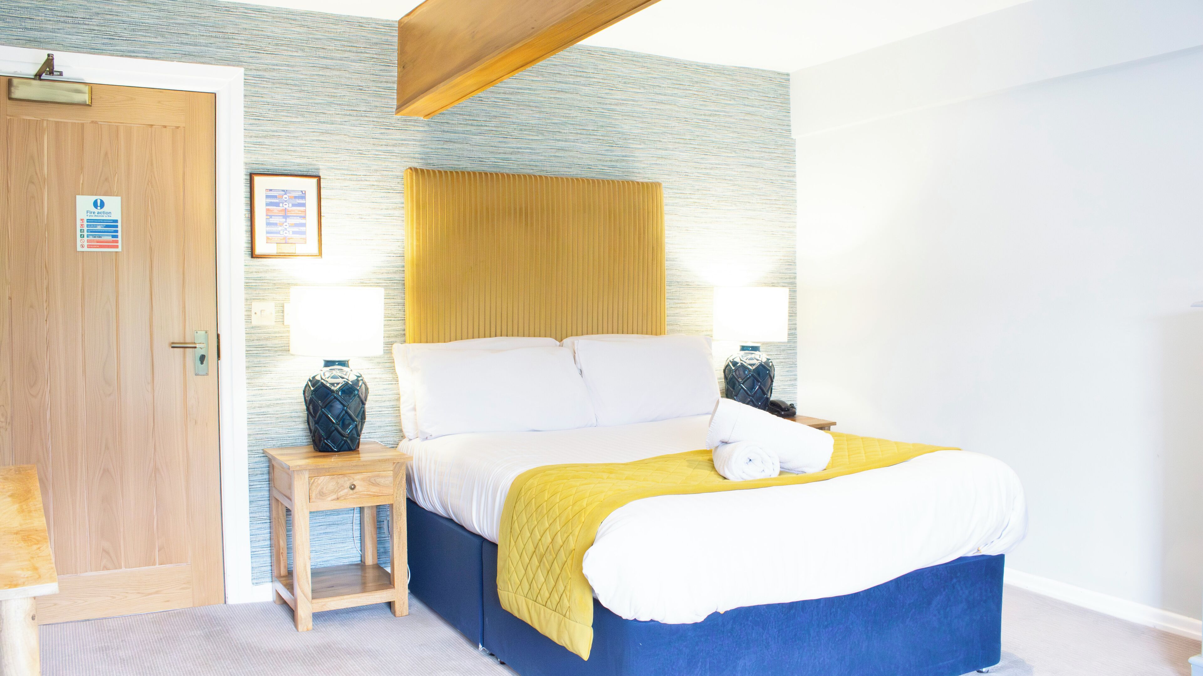 A bedroom with a wooden door, a bedside table with a lamp, a bed with white and yellow bedding, and a blue headboard.