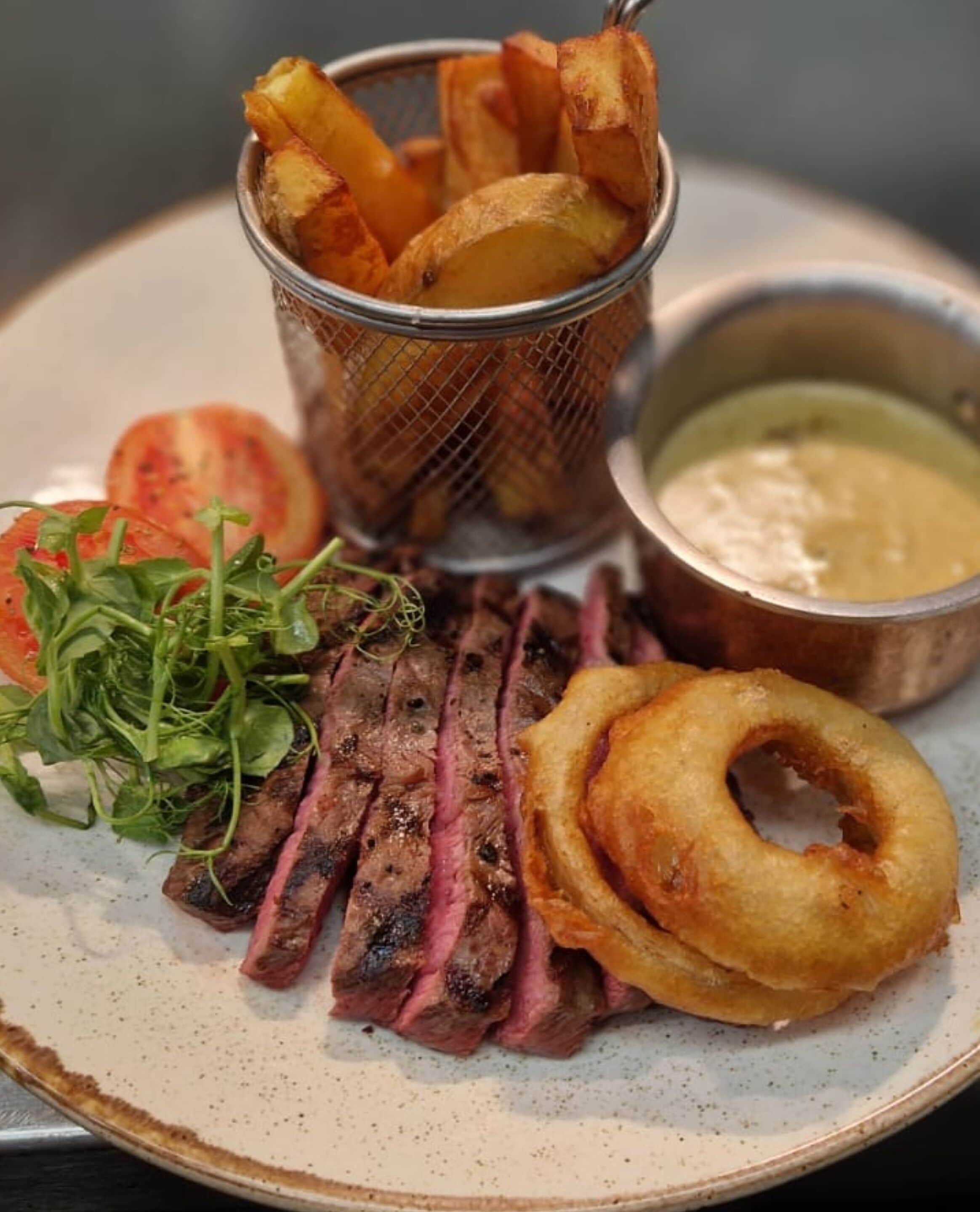 A plate with sliced steak, onion rings, and a small basket of fries, accompanied by a side of greens and a dipping sauce.