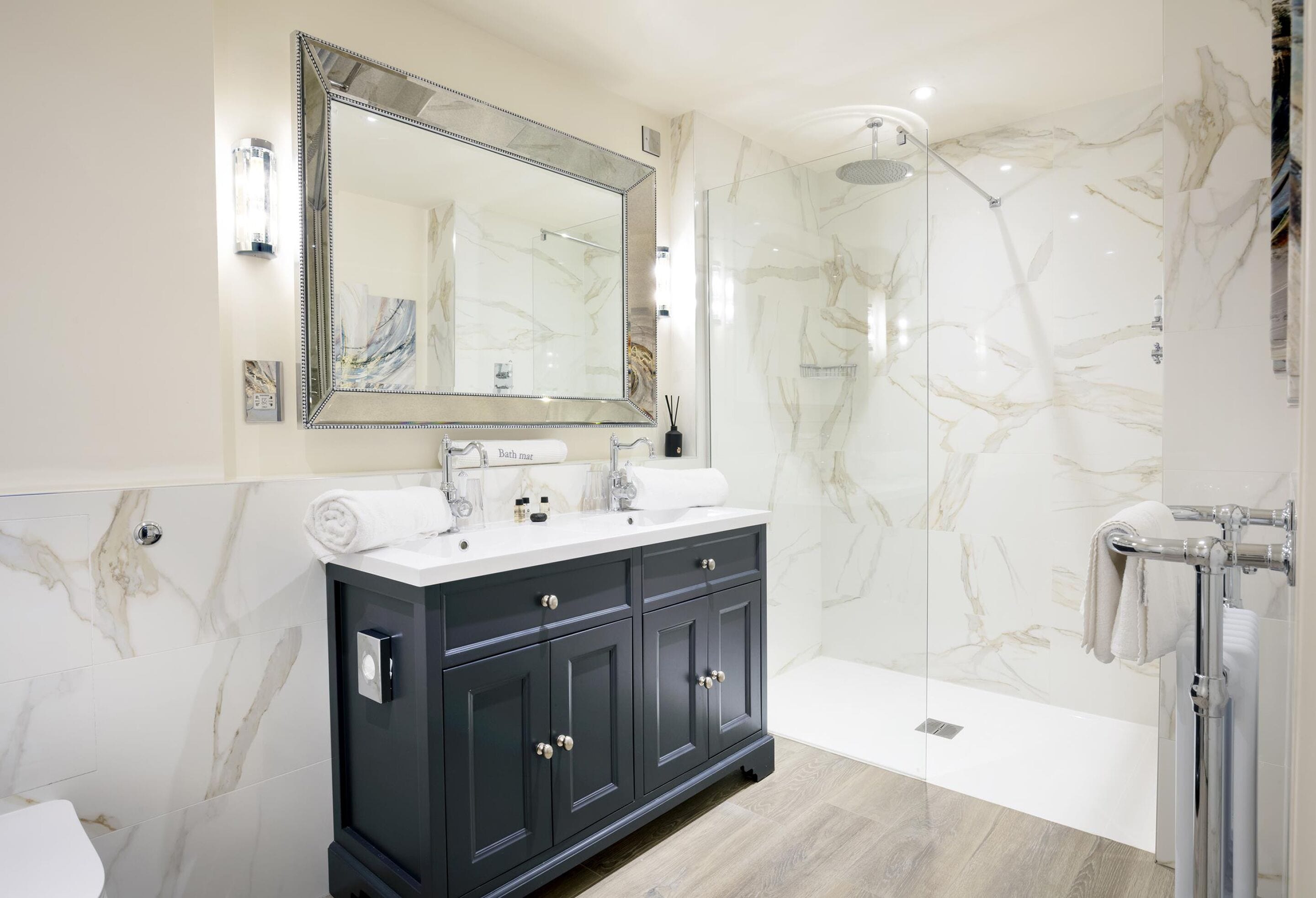 Un baño moderno con ducha de mármol, un espejo grande y un mueble de tocador oscuro con lavamanos.