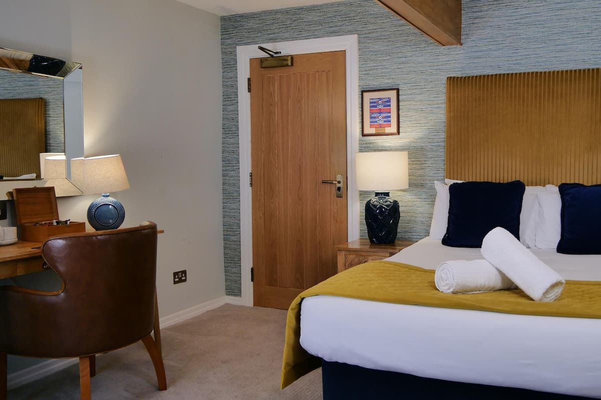 A hotel room with a bed, a desk with a chair, a wooden door, and a framed picture on the wall.