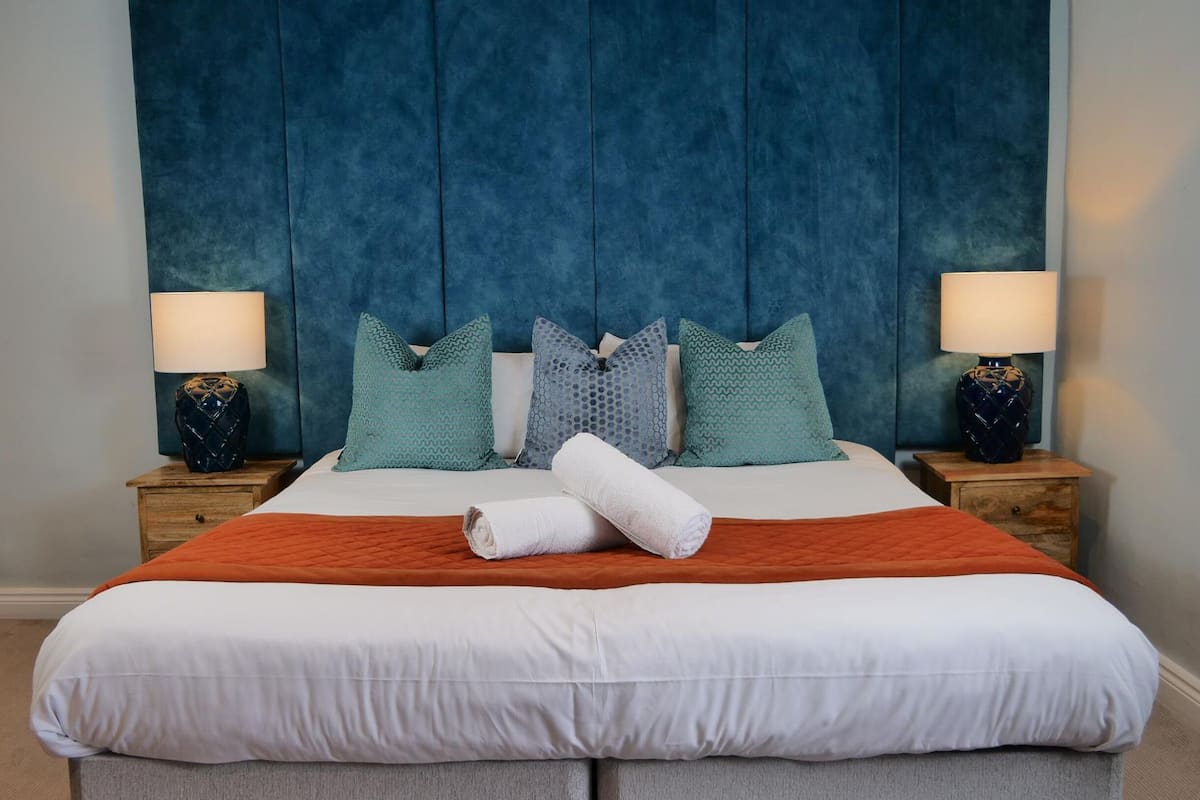A neatly made bed with a white and orange quilt, flanked by two nightstands with lamps, against a blue upholstered headboard.