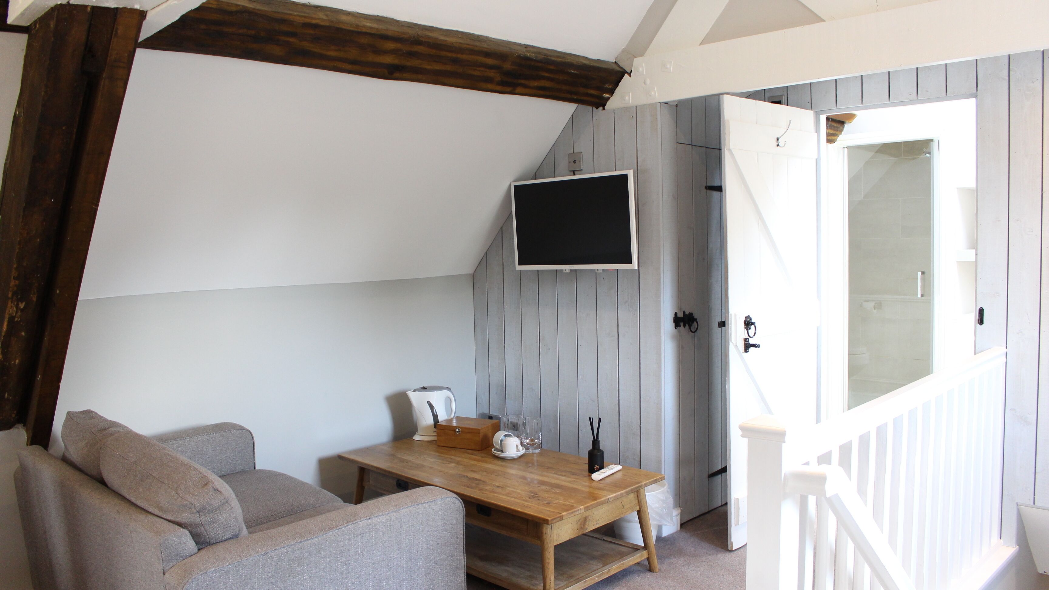 A cozy room with a wooden table, a sofa, a TV mounted on the wall, and a door leading to a bathroom.