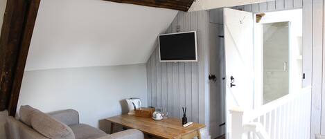 A cozy room with a wooden table, a sofa, a TV mounted on the wall, and a door leading to a bathroom.