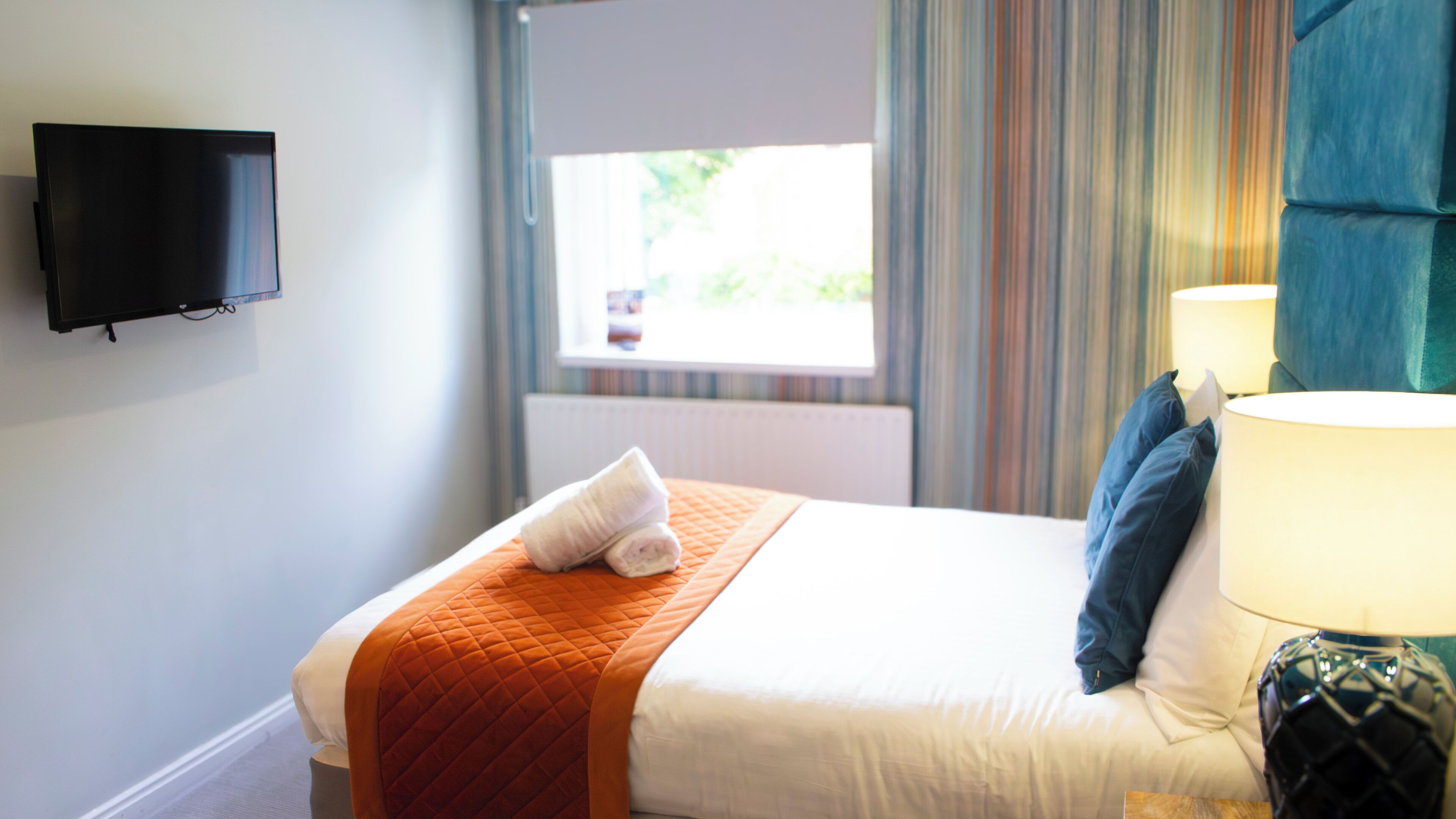 Quarto de hotel com cama, televisão na parede, janela com cortinas e abajur.