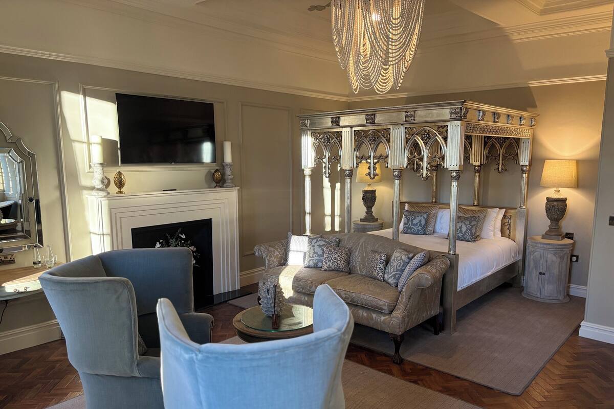 A bedroom with a four-poster bed, a television above the fireplace, and a chandelier.