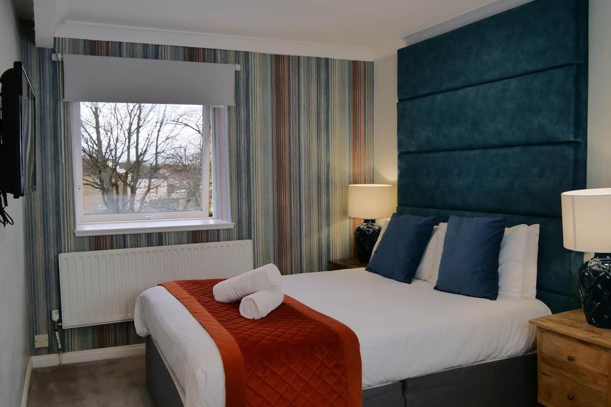 A hotel room with a bed, a window, a radiator, a TV, and a bedside table with lamps.