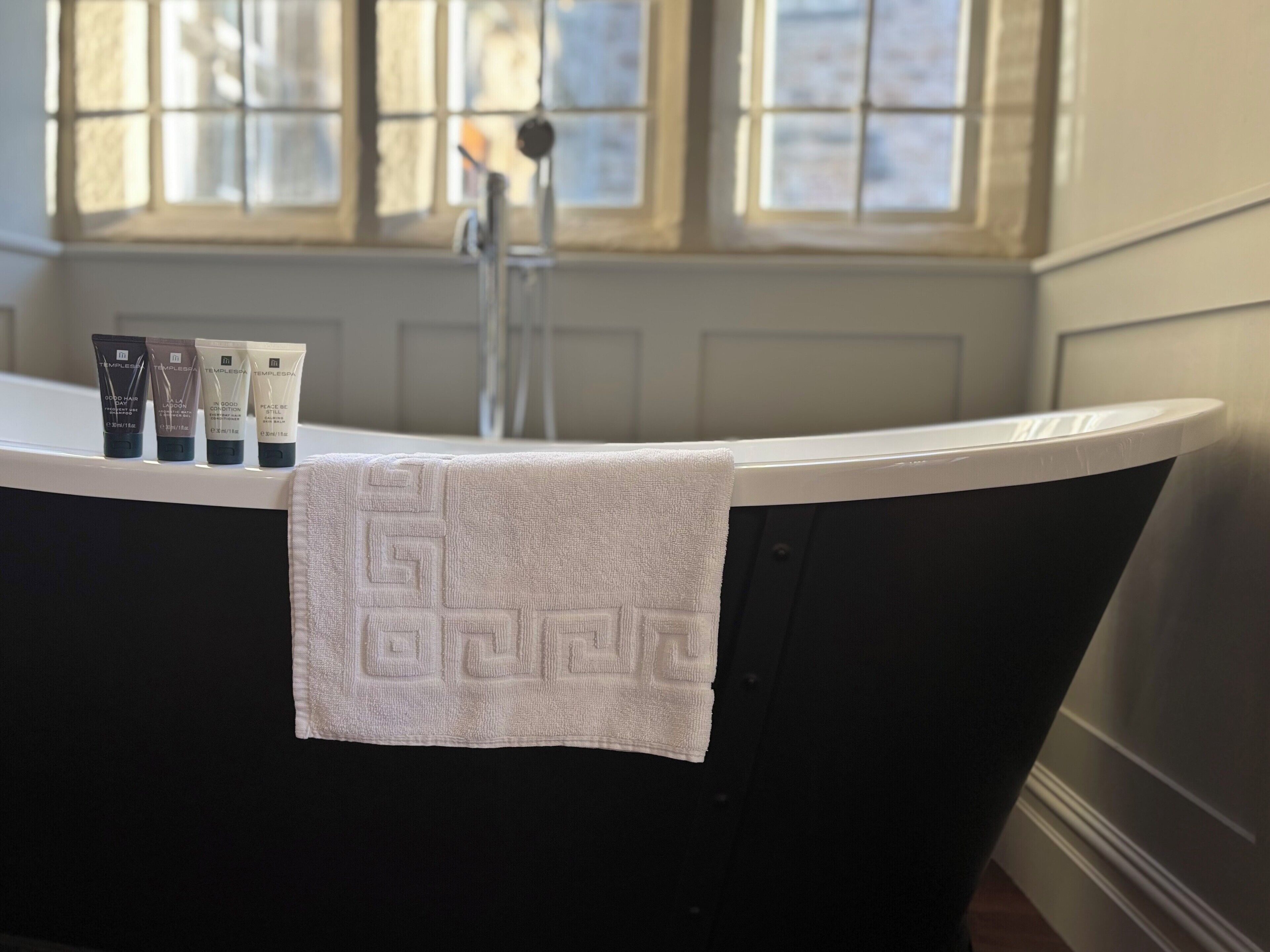 A bathtub with a towel draped over it, a window with a view of buildings, and three skincare products on the edge of the tub.
