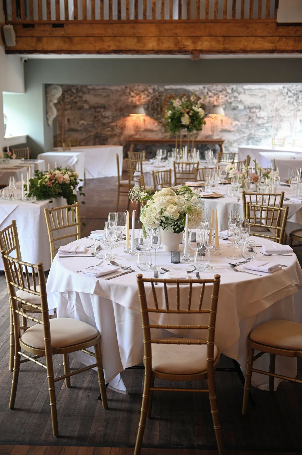 A banquet hall with round tables set for a formal event, featuring white tablecloths, gold chairs, and floral centerpieces.
