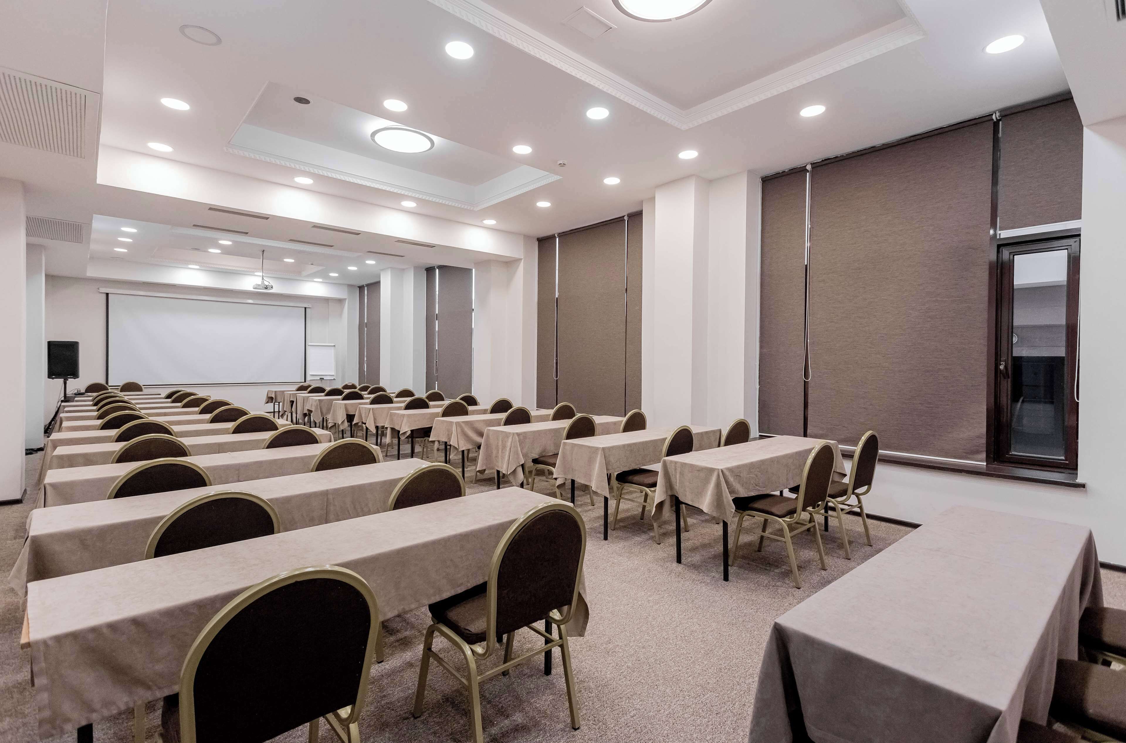 A conference room with multiple tables and chairs, a projector screen, and a large window.