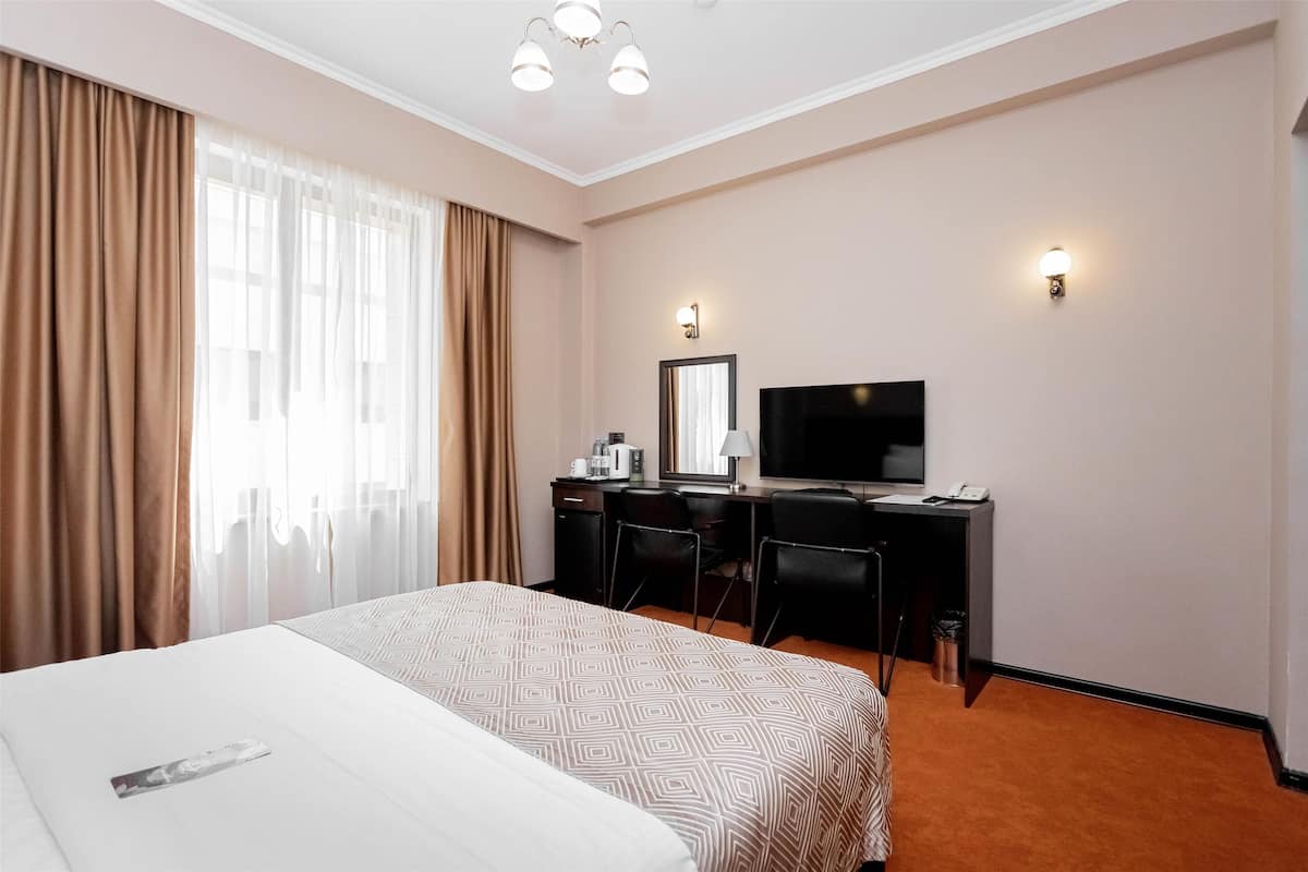A hotel room with a large bed, a desk with a computer, a TV, and a window with curtains.