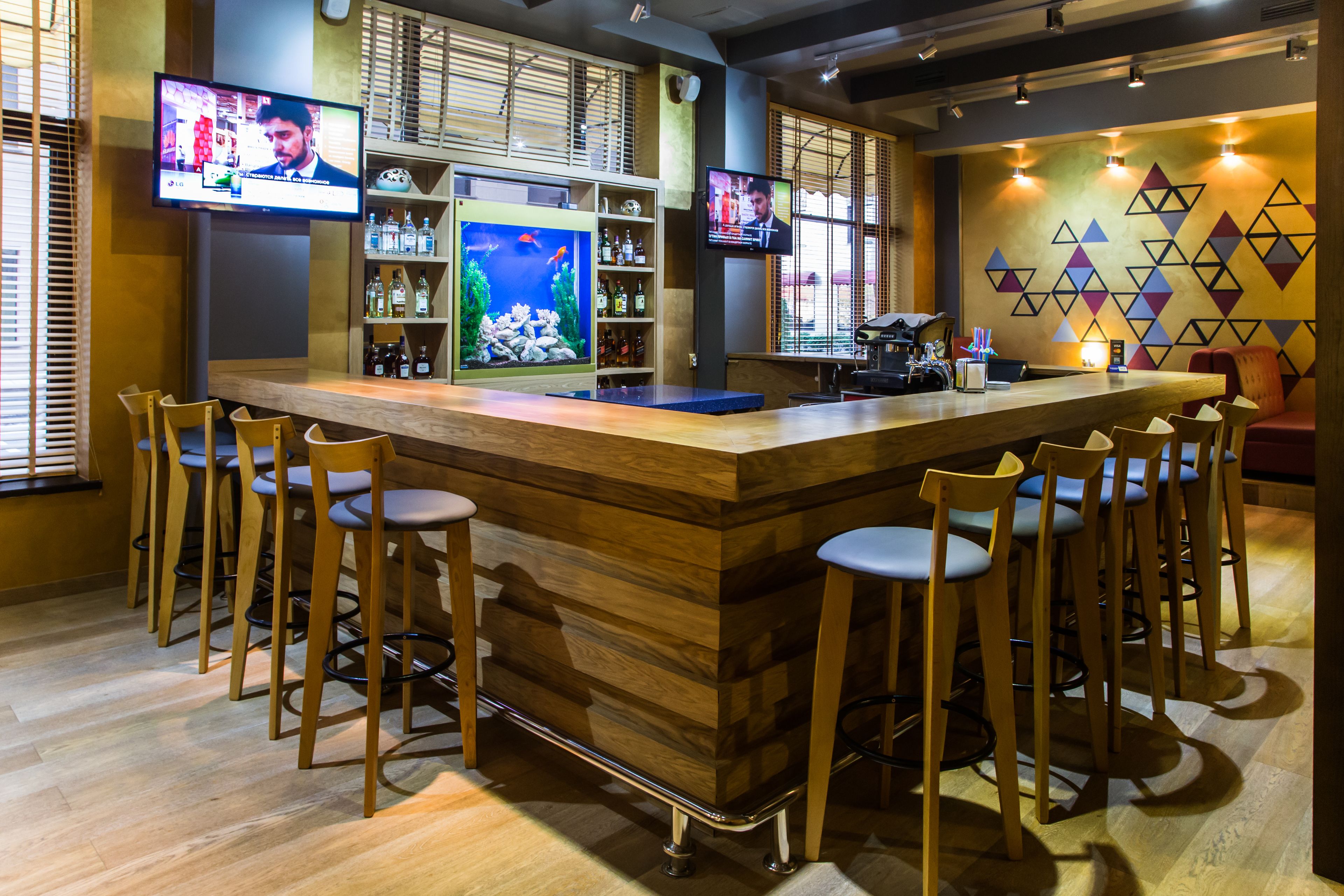 A bar area with a wooden counter, multiple bar stools, and two televisions mounted on the wall.