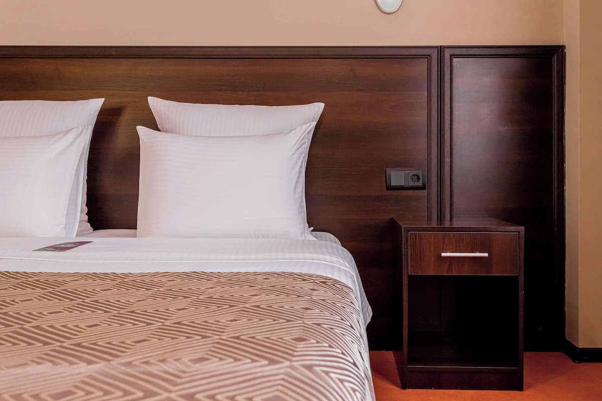 A neatly made bed with a patterned bedspread and two pillows, a small bedside table with a drawer, and a wall-mounted light fixture.