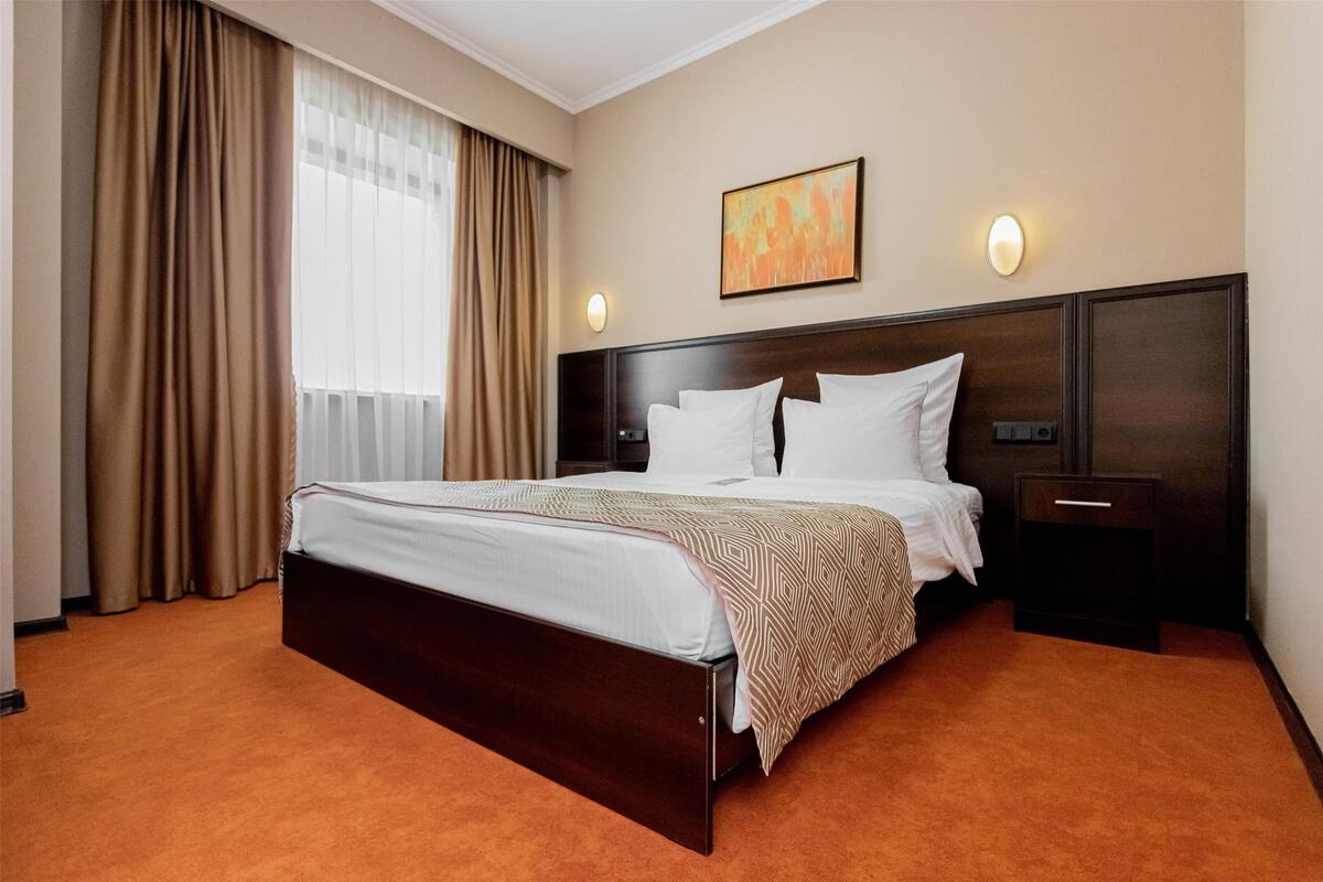 A hotel room with a large bed, a nightstand, a wall-mounted light, and a window with curtains.