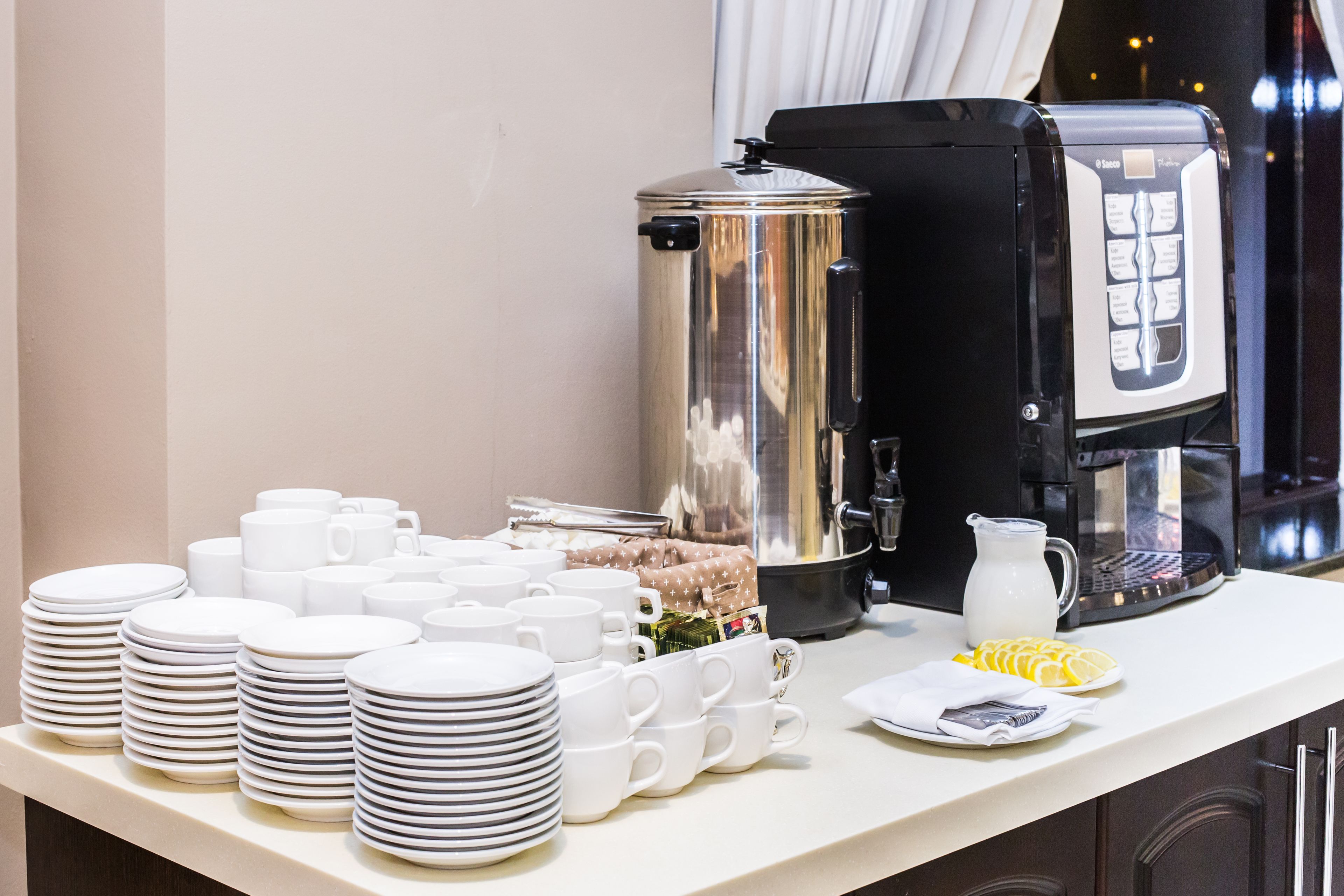 Een buffetopstelling met stapels witte borden en bekers, een zilveren voedselcontainer, een koffiezetapparaat en een melkkruik op een aanrecht.