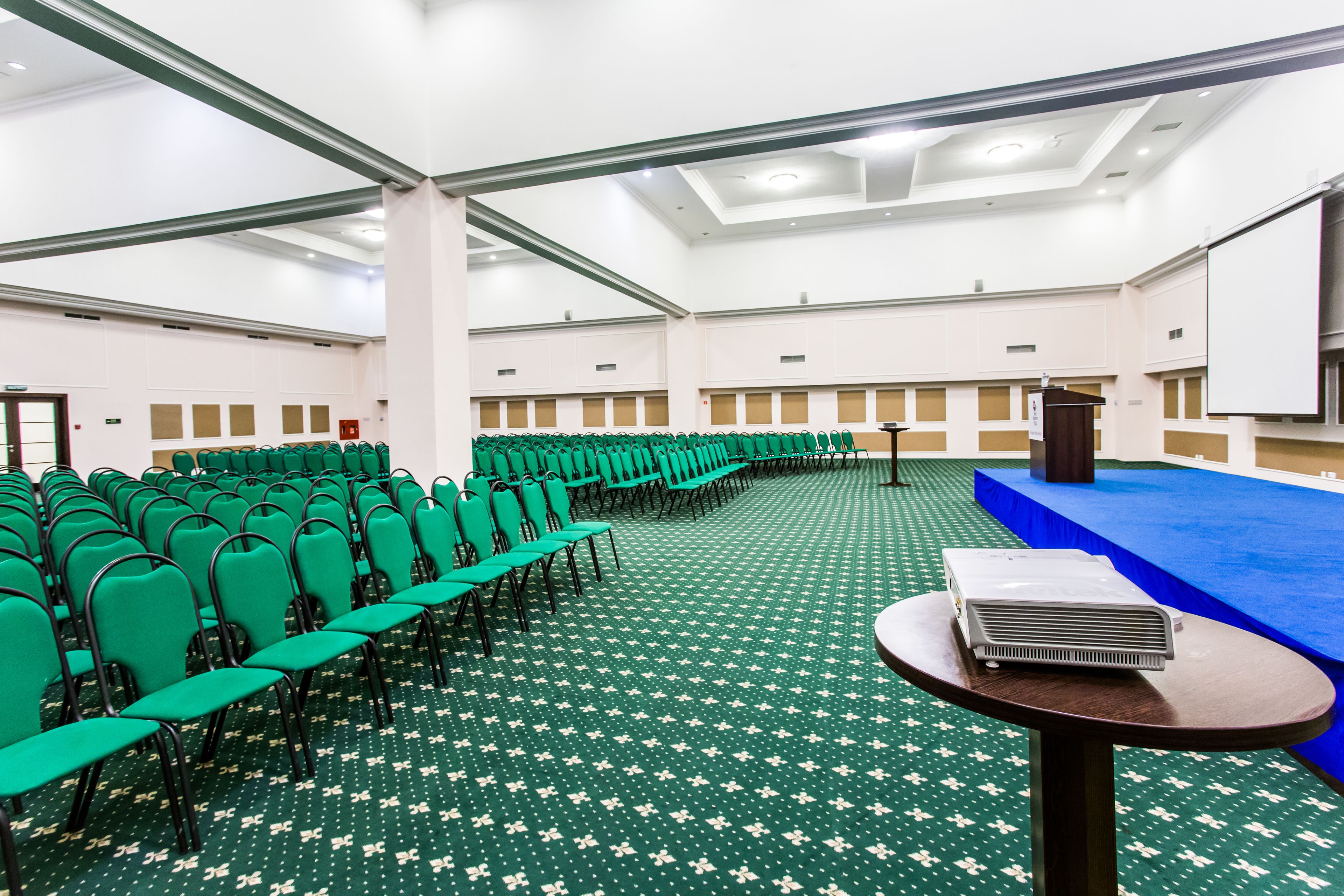 A conference room with rows of green chairs, a blue stage, and a projector on a table.