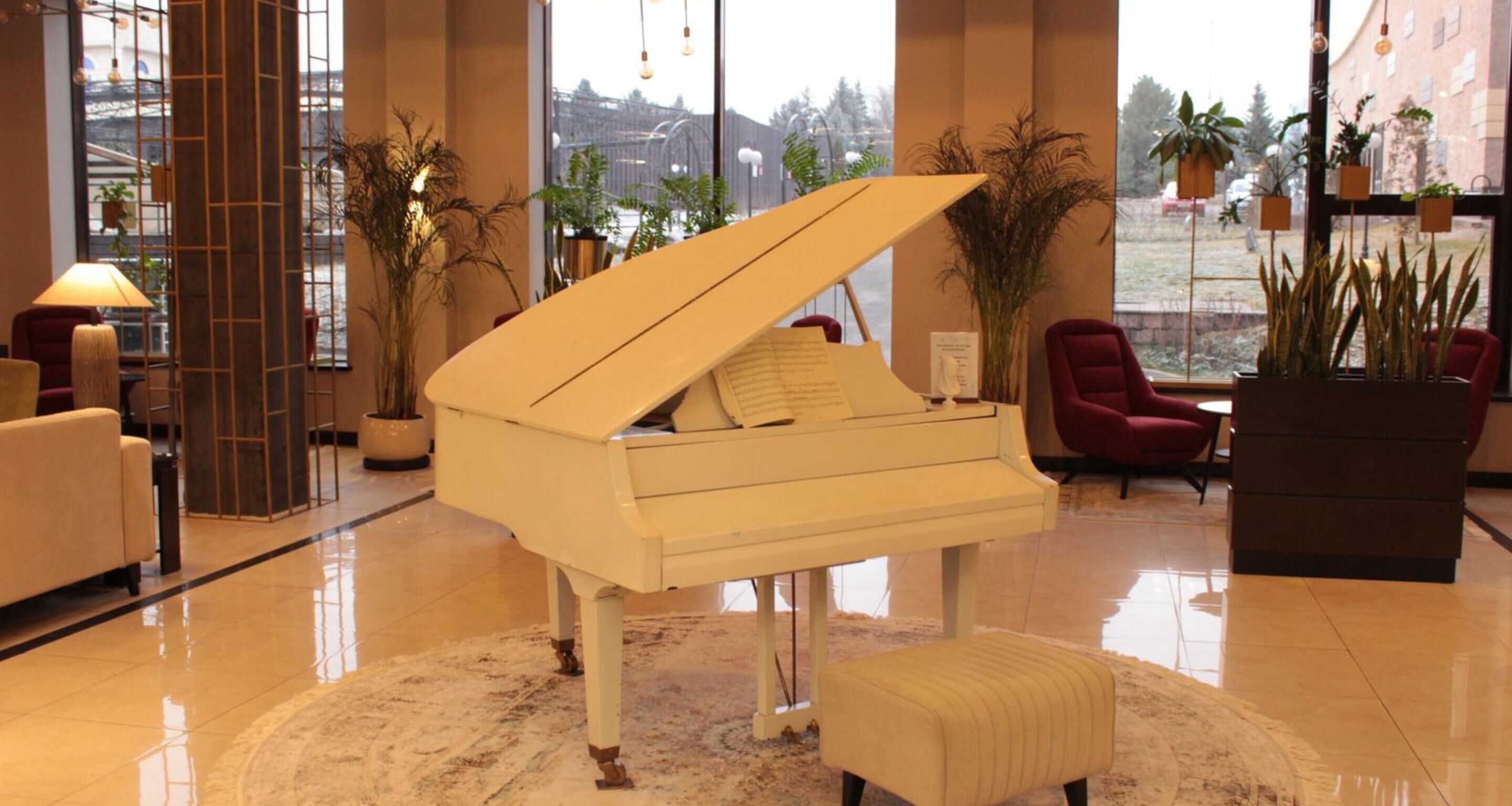 Un piano de cola en un vestíbulo amplio con piso de mármol, sillas rojas y plantas en macetas.